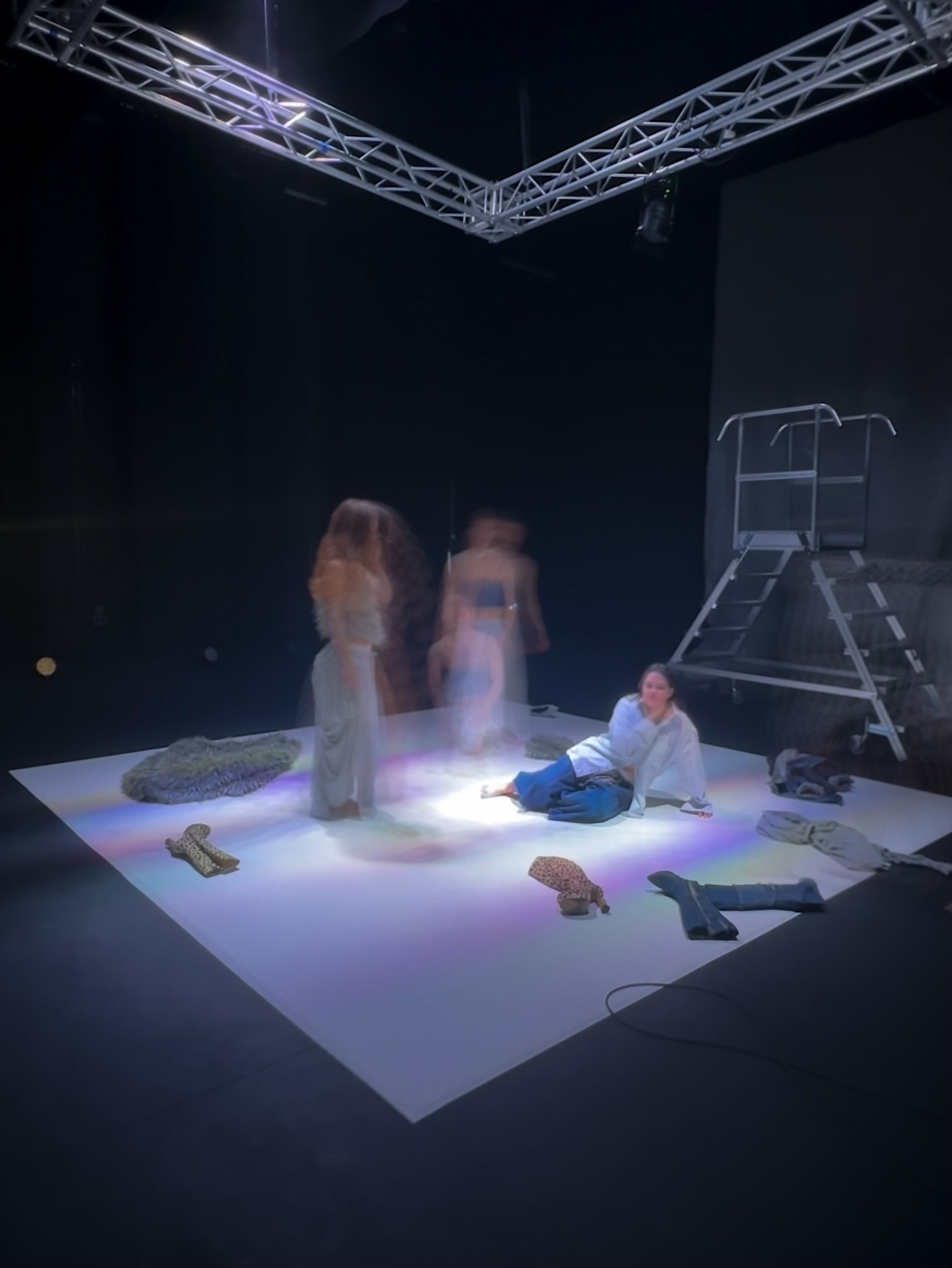 Cuatro personas en un espacio oscuro, con objetos dispersos en el suelo y una estructura metálica en el techo. Una persona está sentada en el suelo, otra de pie y las otras parecen estar en movimiento.
