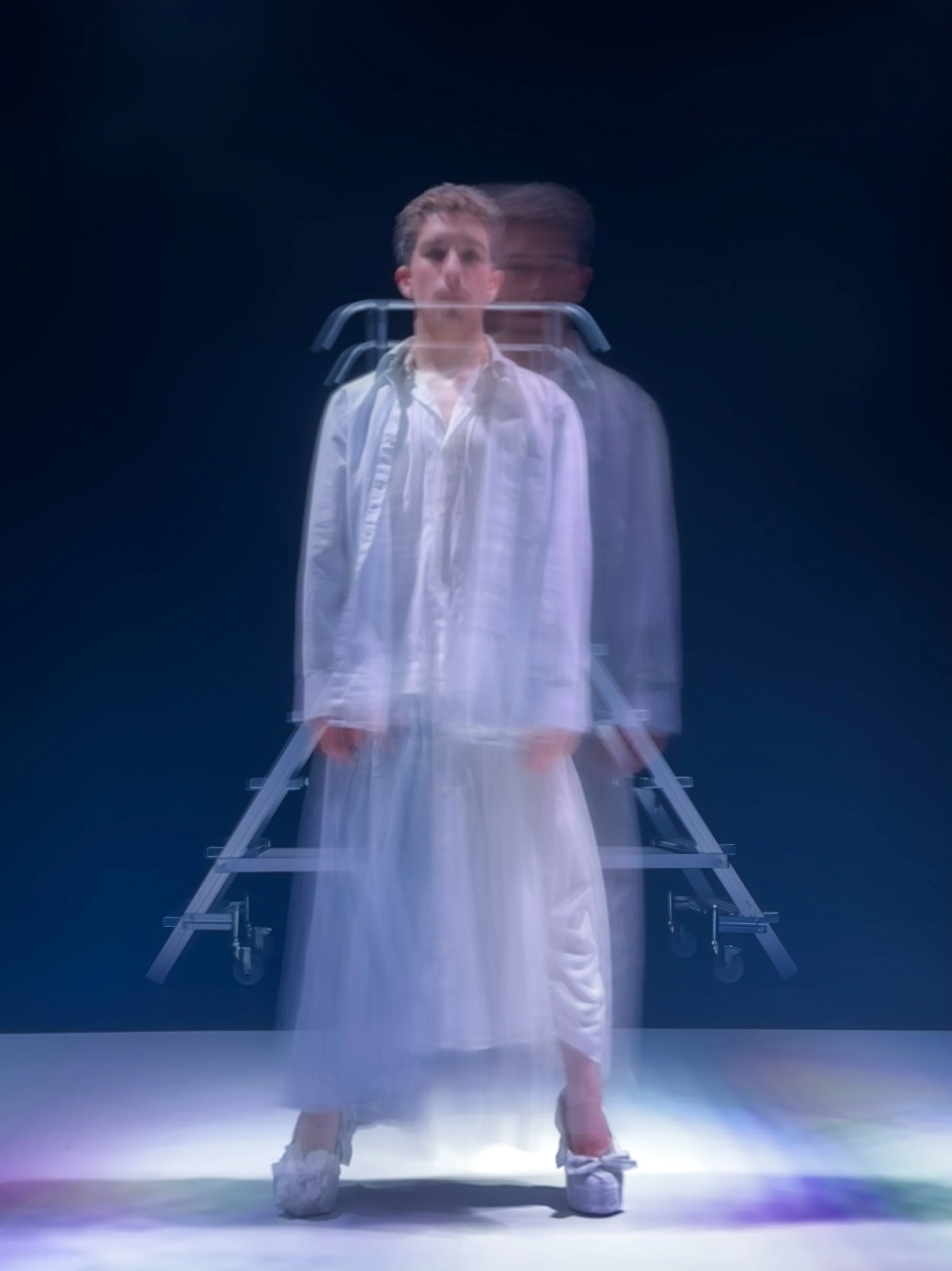Una persona de cabello castaño, vestida con ropa blanca, con una doble exposición que muestra a otra persona con gafas y cabello oscuro, en un fondo oscuro. Está de pie frente a una estructura metálica con ruedas y parece estar en un escenario con il