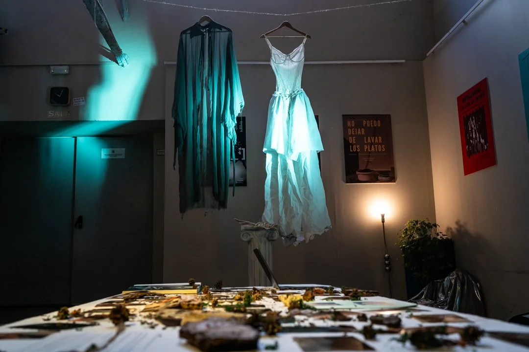 Dos vestidos blancos colgados en perchas en la pared, con un fondo con señalización y una mesa con objetos en primer plano.