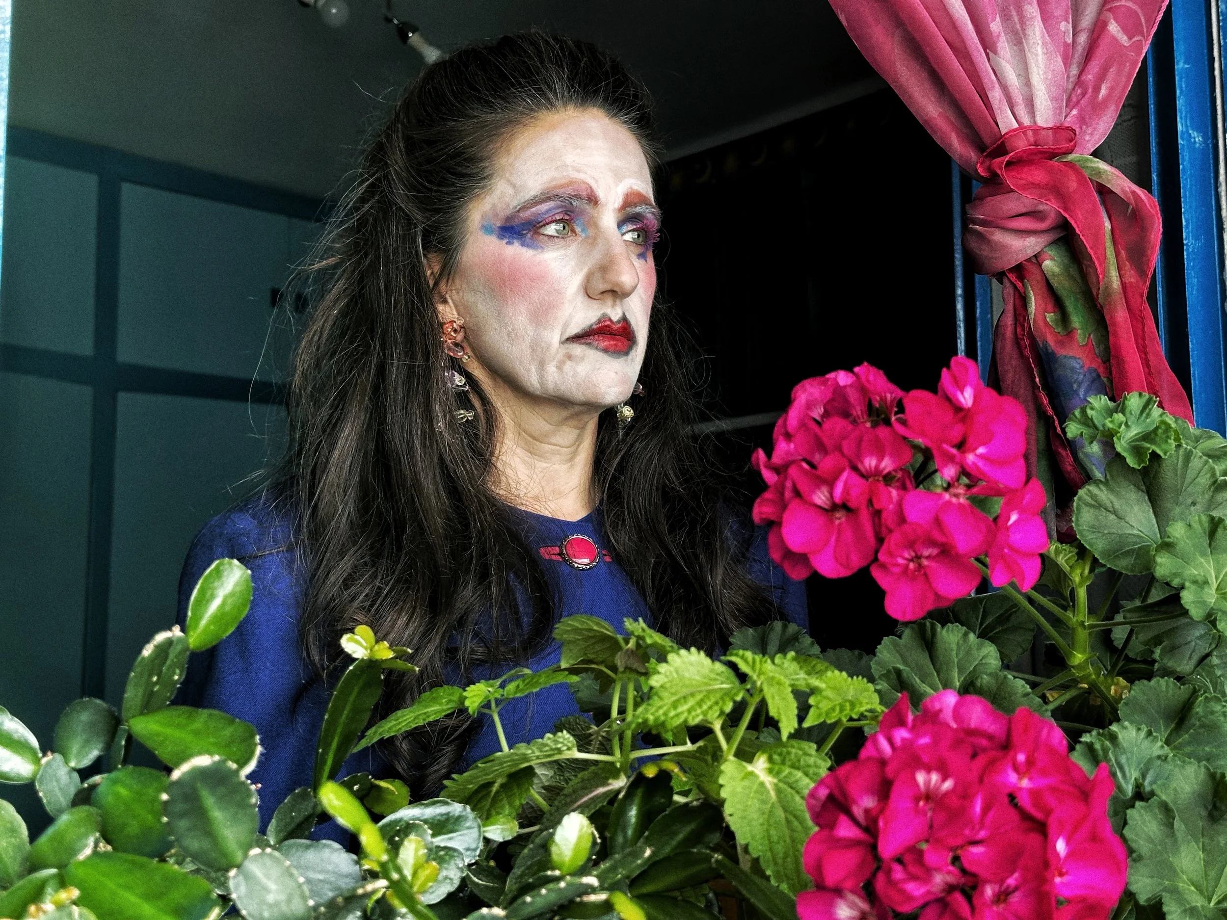 Una mujer con maquillaje artístico y expresión pensativa, rodeada de plantas y flores de colores vivos.