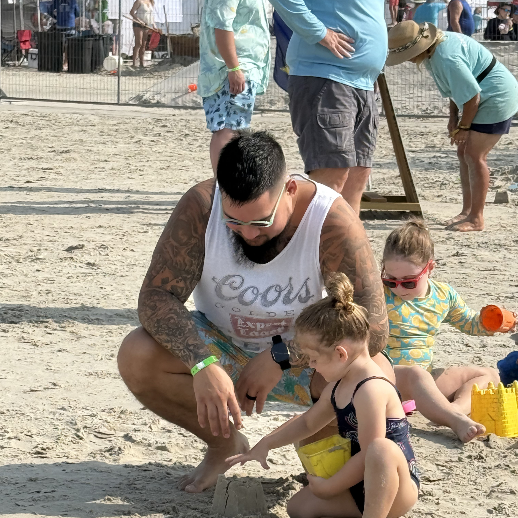 A man with tattoos and sunglasses crouching on a sandy beach playing with two young girls, one in a swimsuit with her hair in a bun and the other in a yellow patterned outfit with sunglasses. People are in the background, some standing, some bending down, on a sunny beach.