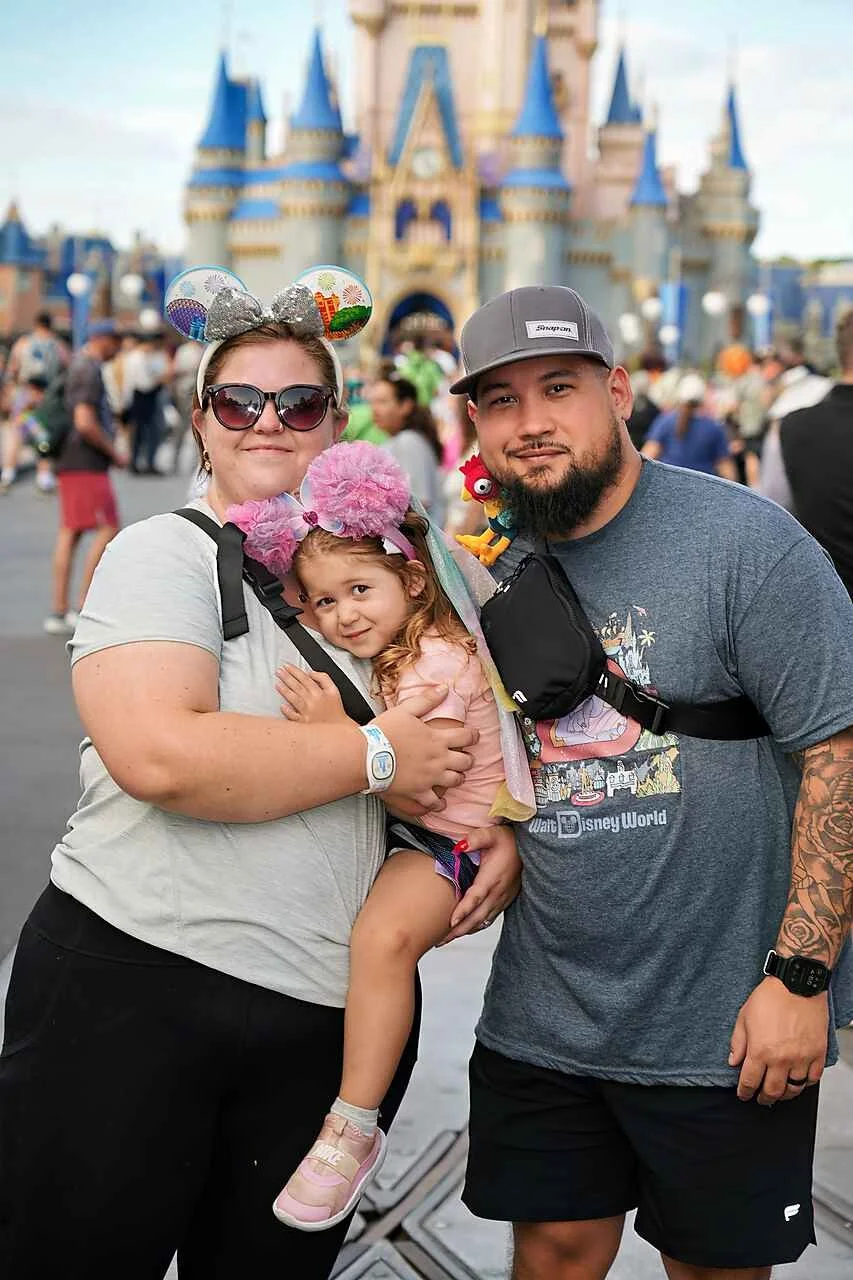 Family posing in front of Disneyland castle, wearing Disney-themed accessories, with many visitors in the background.