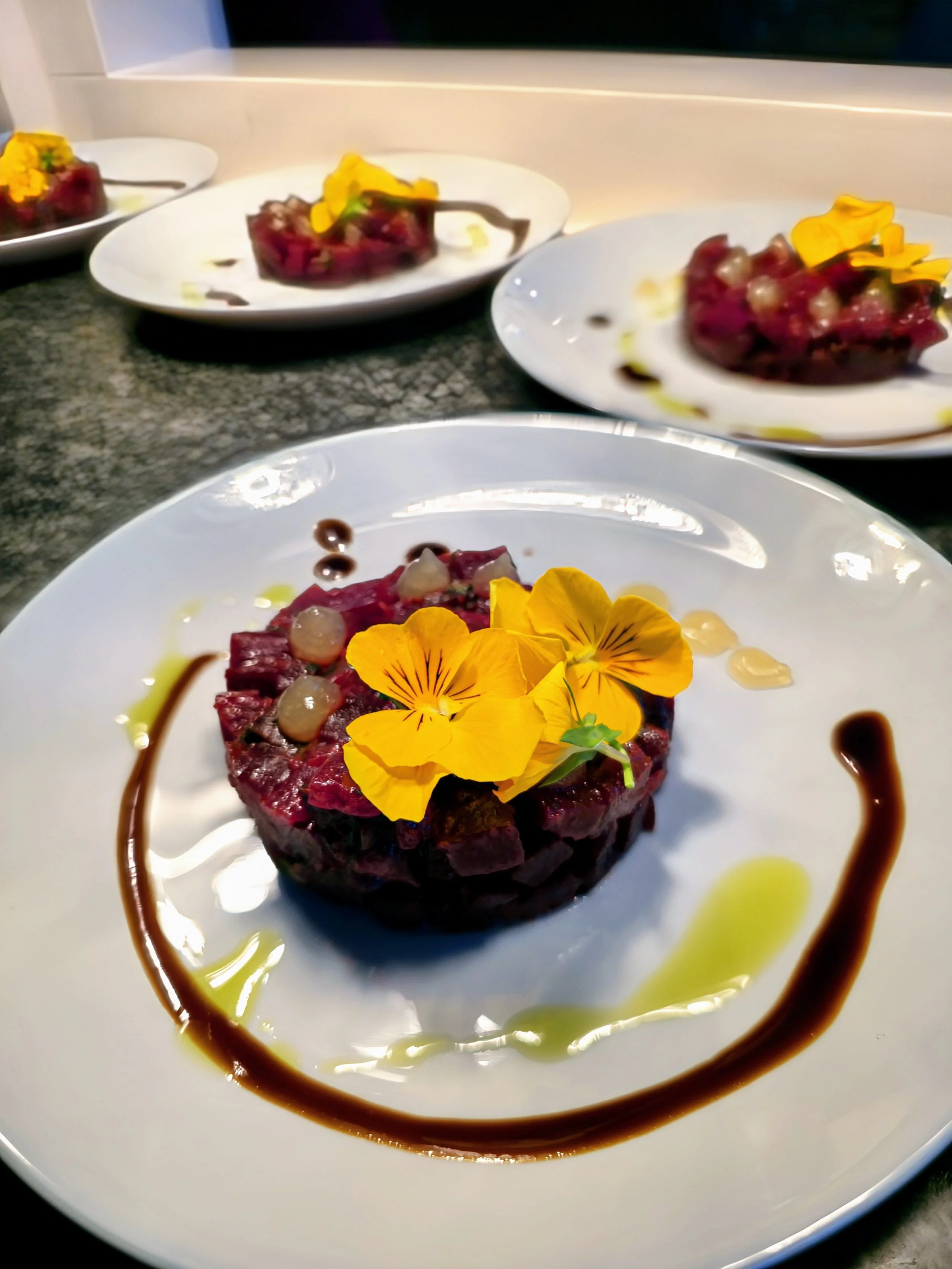 A plate of raw beef tartare topped with yellow edible flowers, pearl onions, and garnished with olive oil and balsamic glaze on a white plate.