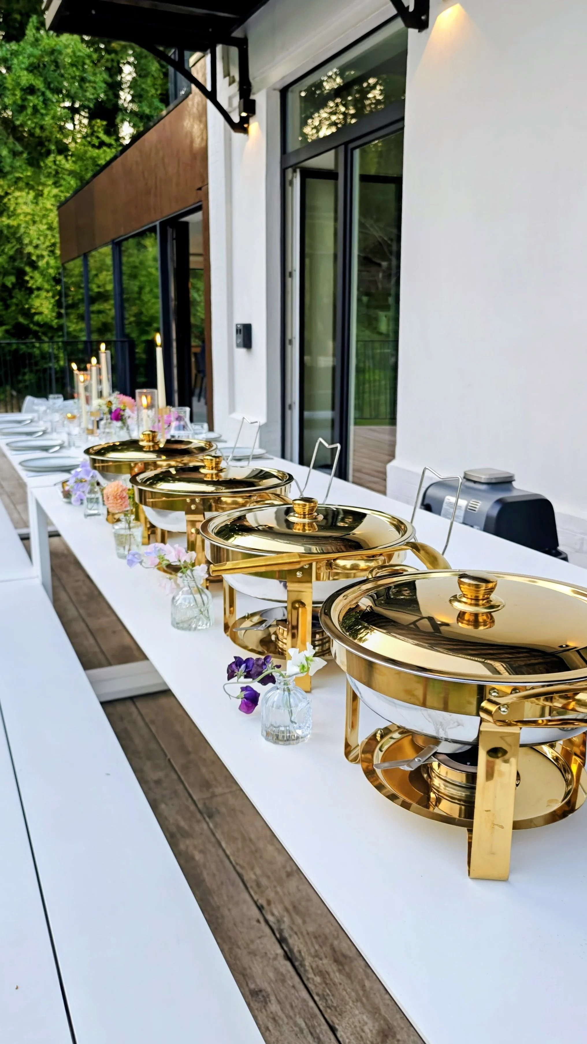 A long outdoor dining table set with gold chafing dishes, small vases of colorful flowers, and lit candles, located on a patio next to a modern house with large glass doors and windows, surrounded by trees.