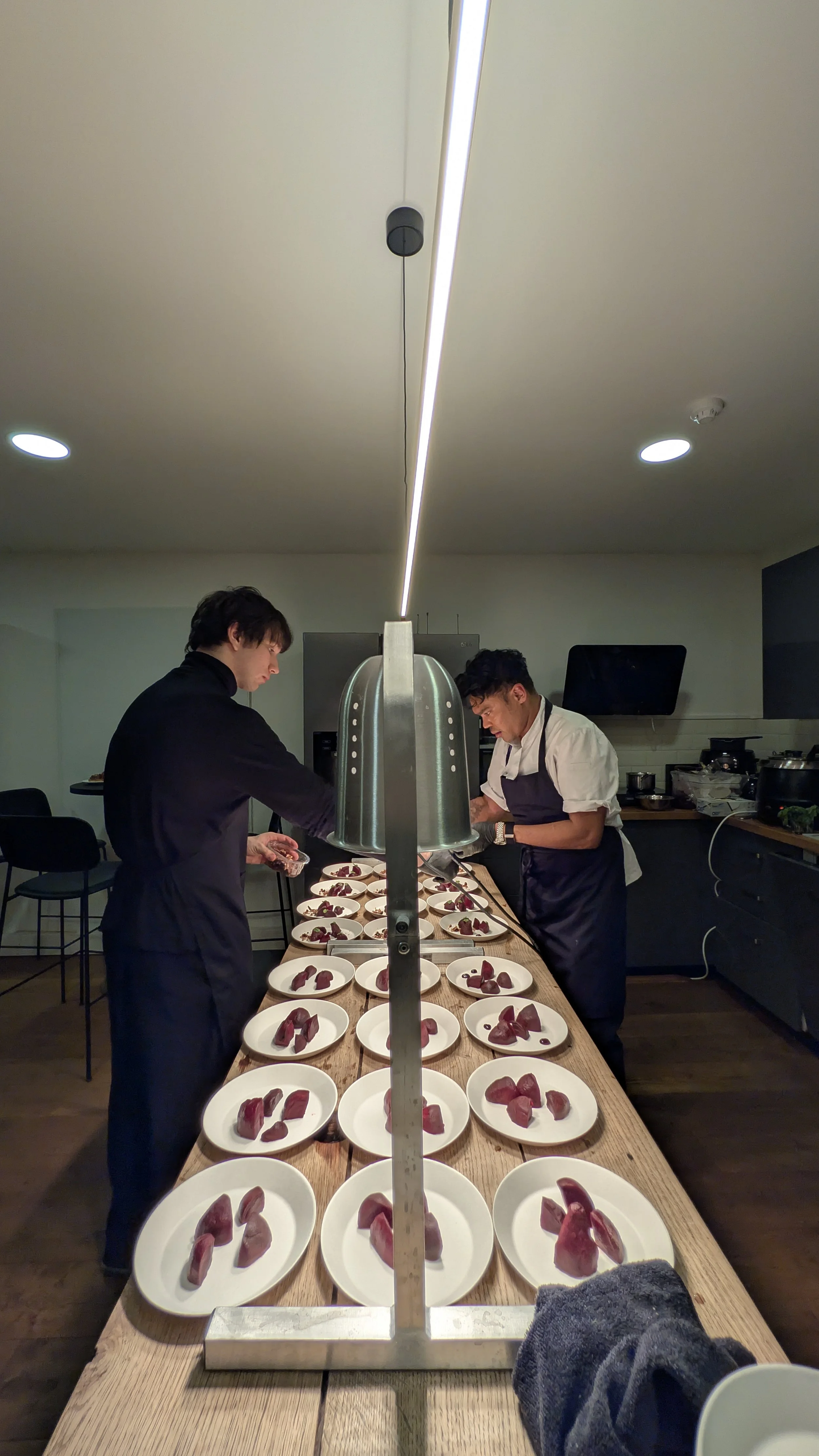 Two chefs preparing multiple plates of raw meat slices in a professional kitchen.