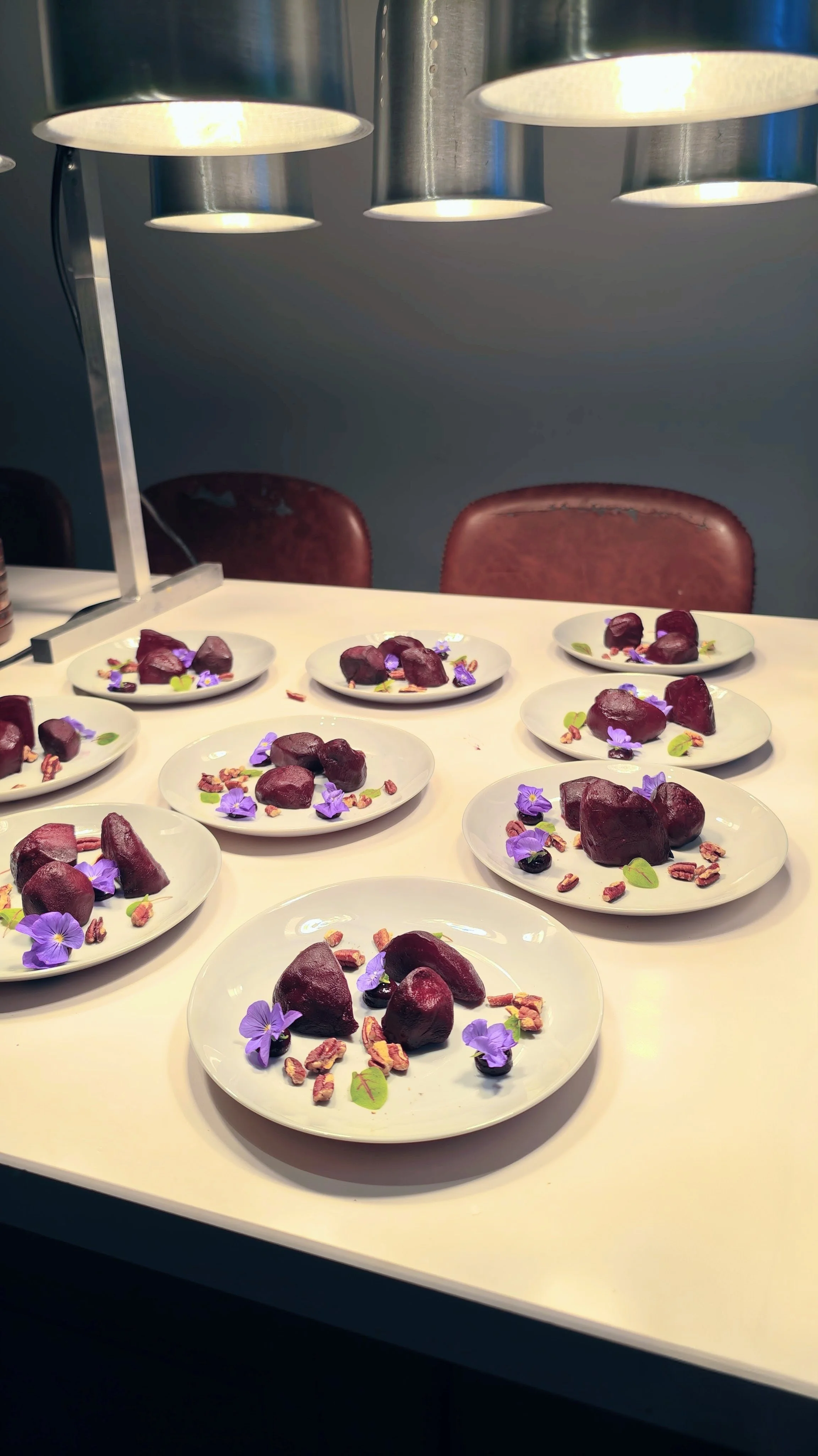 Multiple white plates each with purple-colored beets, purple flowers, chopped pecans, and small green leaves, arranged on a white table under overhead lighting.