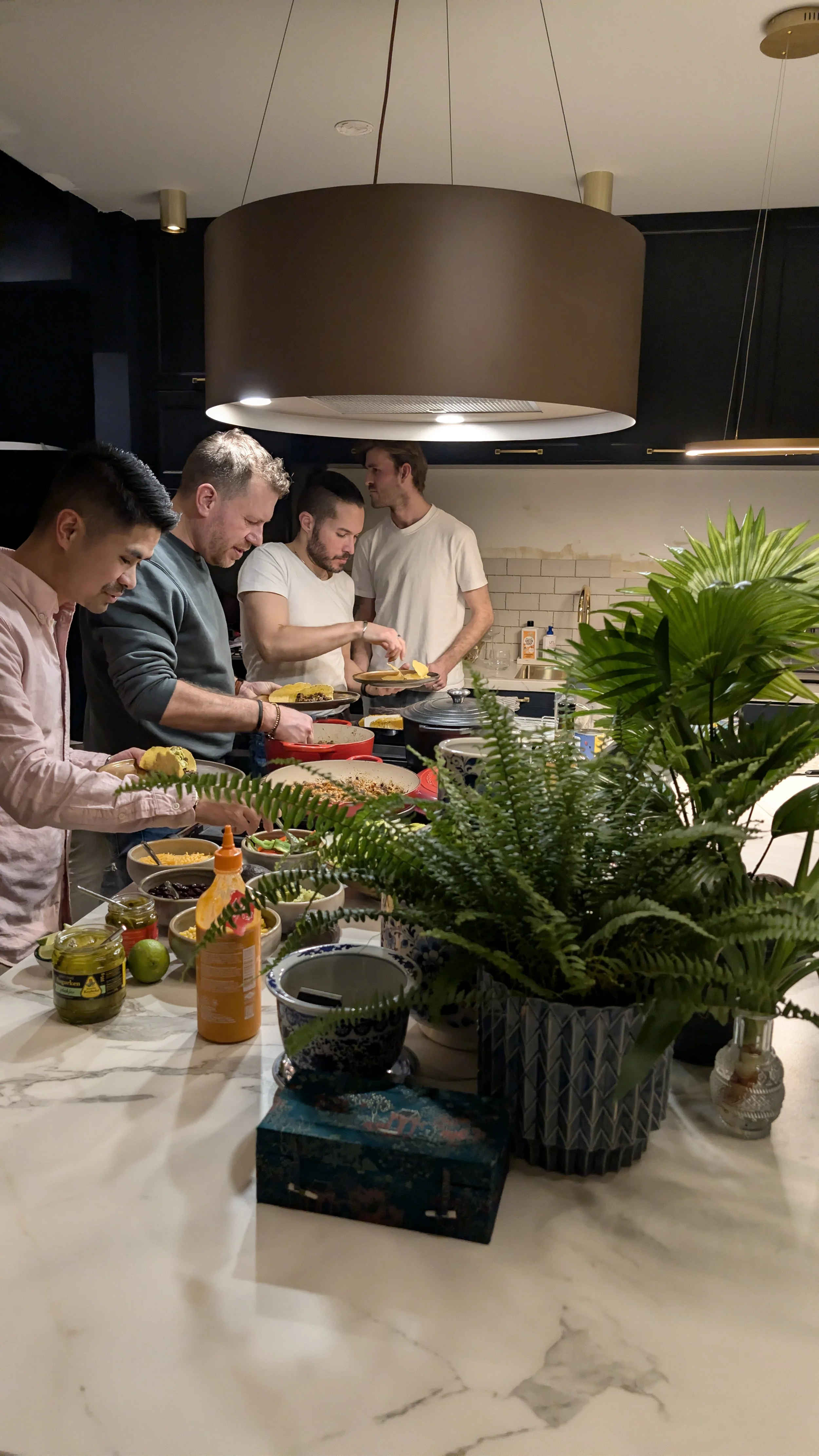 Group of five men preparing food in a modern kitchen, with a table decorated with potted plants, condiments, and bowls of food.