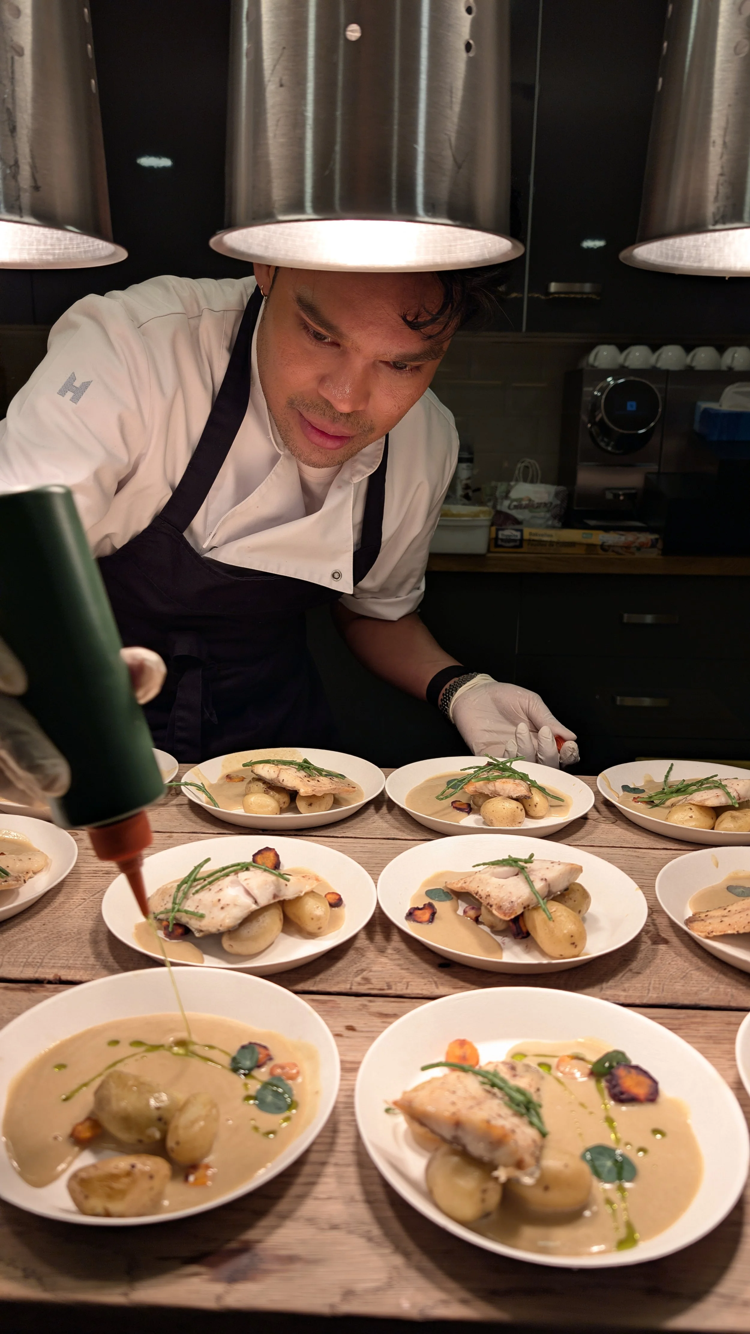 Chef garnishing plates of fish with sauce, potatoes, and greens in a professional kitchen.