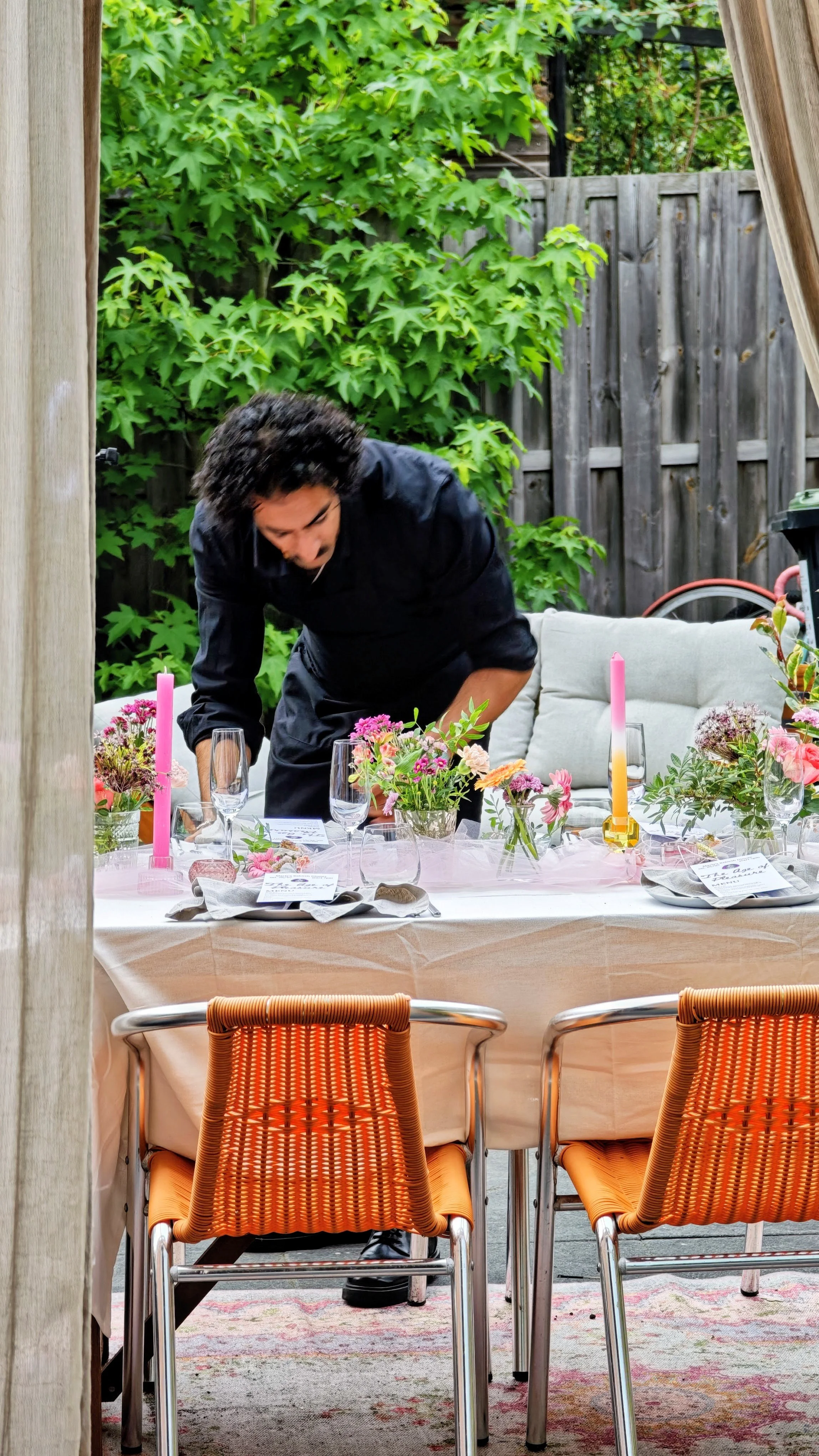A woman setting an elegant outdoor table decorated with pink flowers, tall pink candles, and place settings, with lush green foliage in the background.