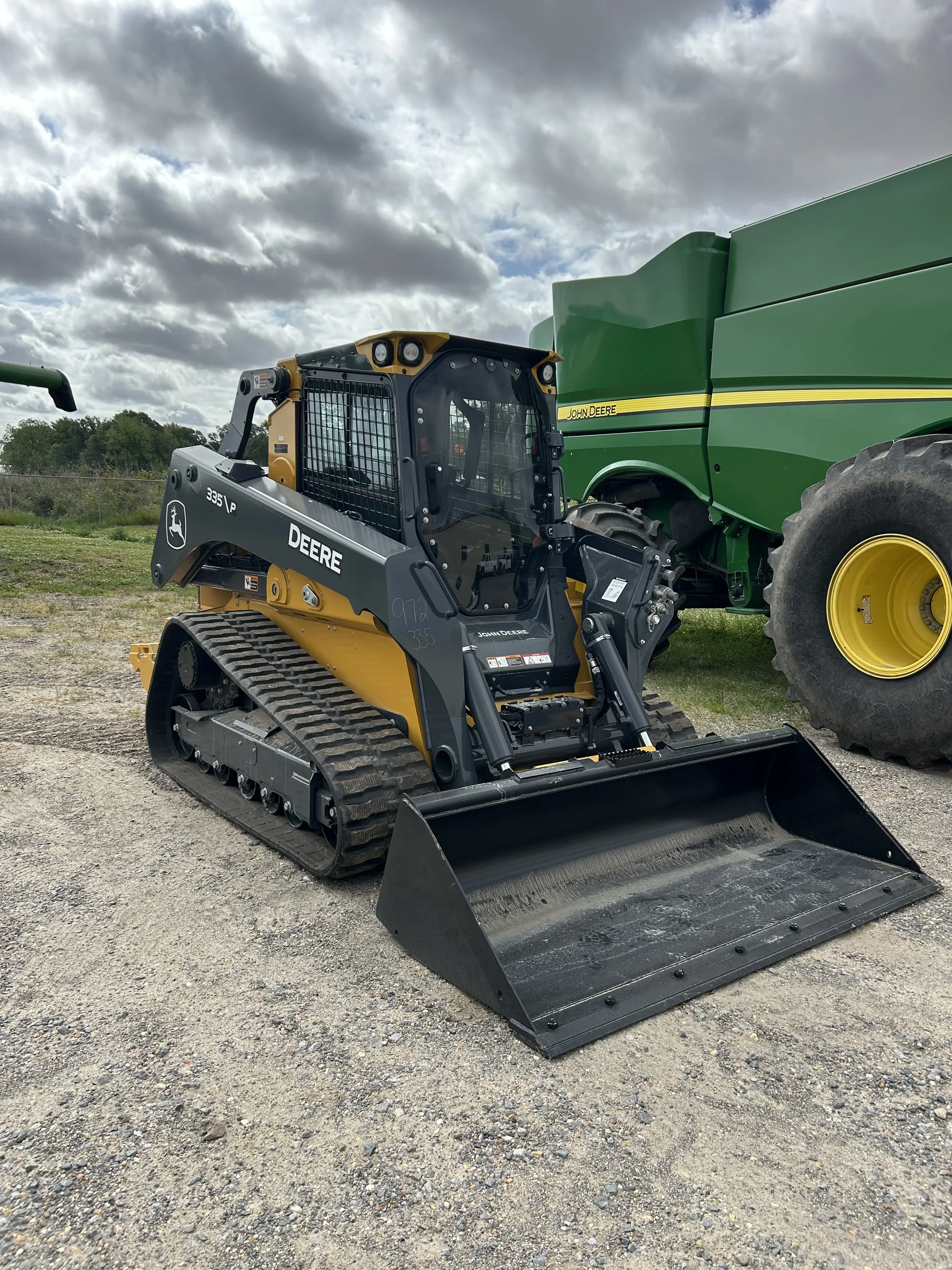 A new John Deere compact track loader with a front bucket attachment parked outdoors on a gravel surface under a cloudy sky.