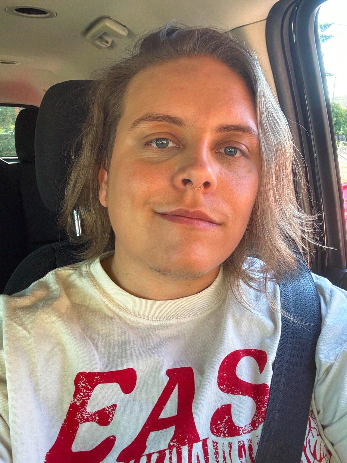 A person with shoulder-length hair, wearing a nose ring, sitting in a car with a seatbelt on, and wearing a white t-shirt with red text.