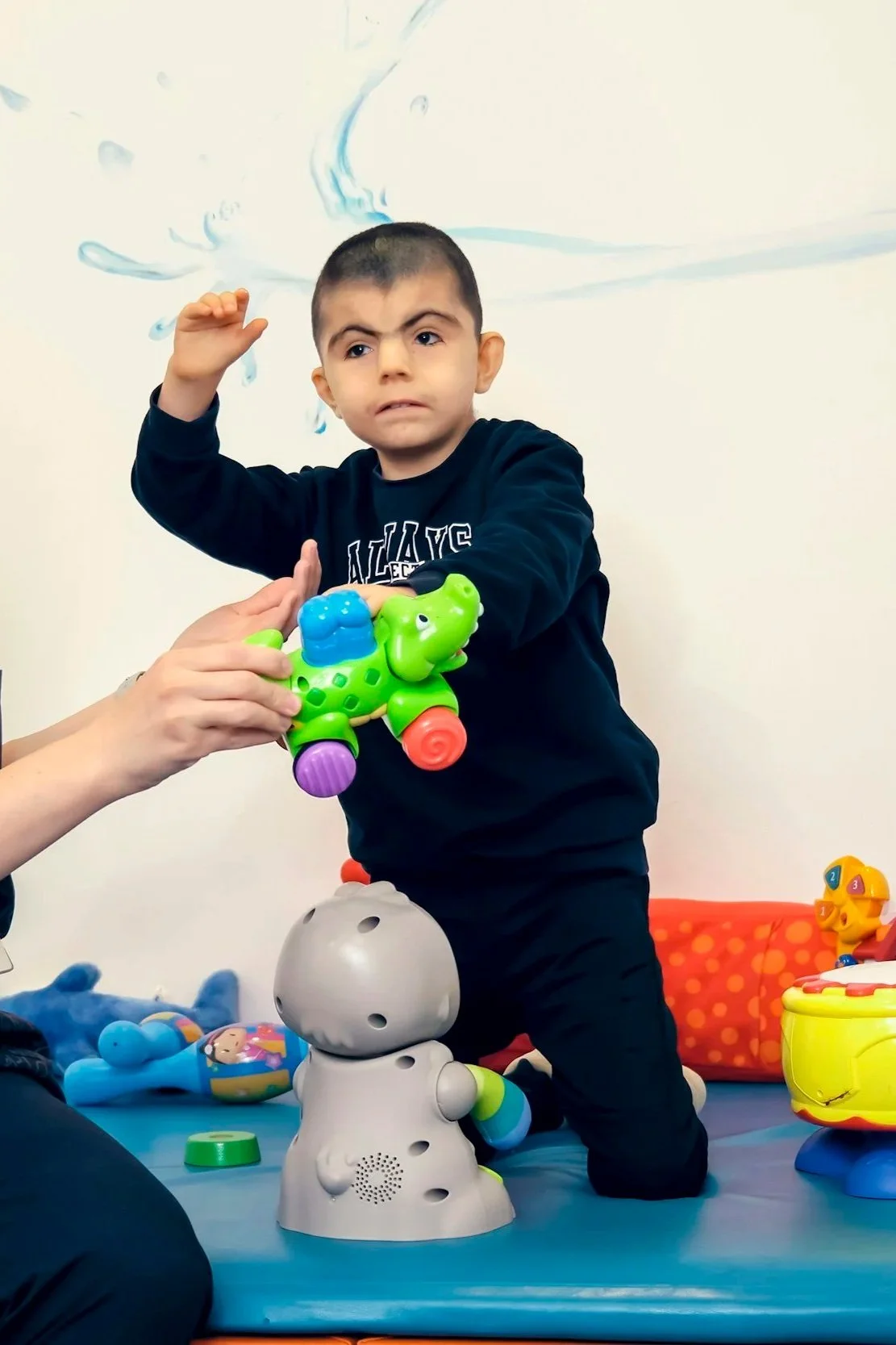 A young boy with short dark hair and a serious expression playing with colorful toys, including a green dinosaur toy, in a playroom. An adult's hand is holding the dinosaur toy in front of him. There are other toys and a red polka-dotted container in the background.
