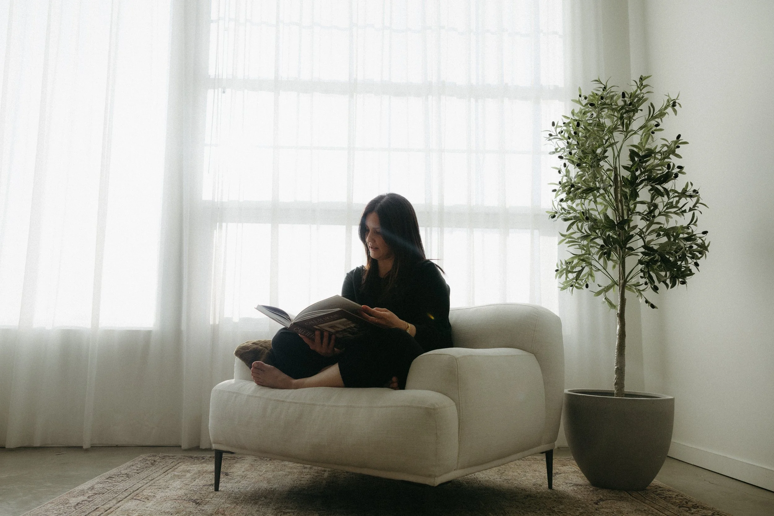 Woman sitting on a cream-colored armchair reading a magazine in a room with large windows and sheer curtains, with a tall potted plant nearby.