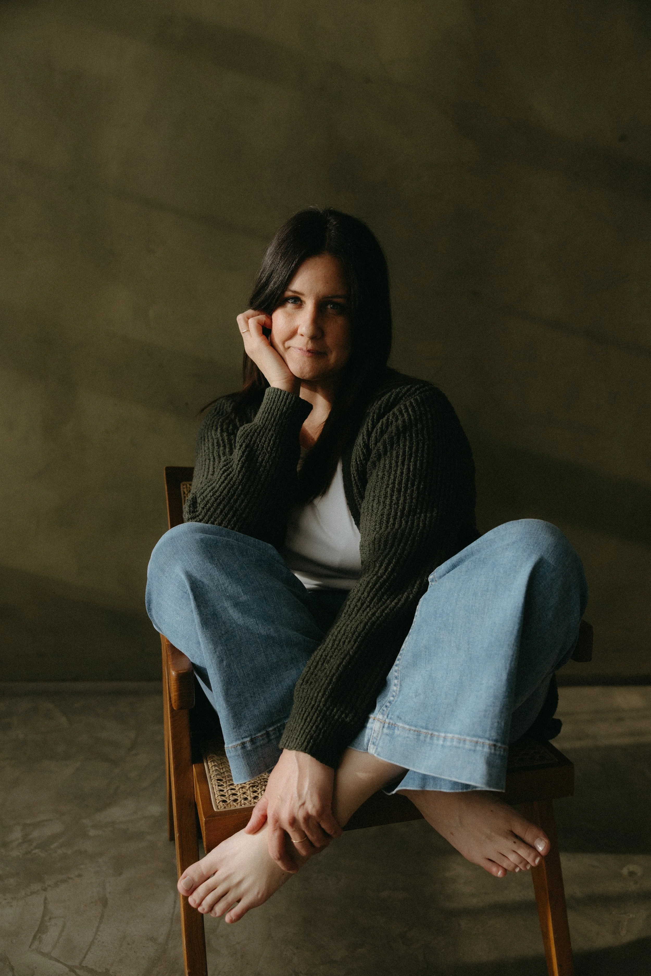 A woman with dark hair sitting on a chair with her legs crossed, wearing a green sweater and blue jeans, smiling with her hand on her face in an indoor setting with a dark background.