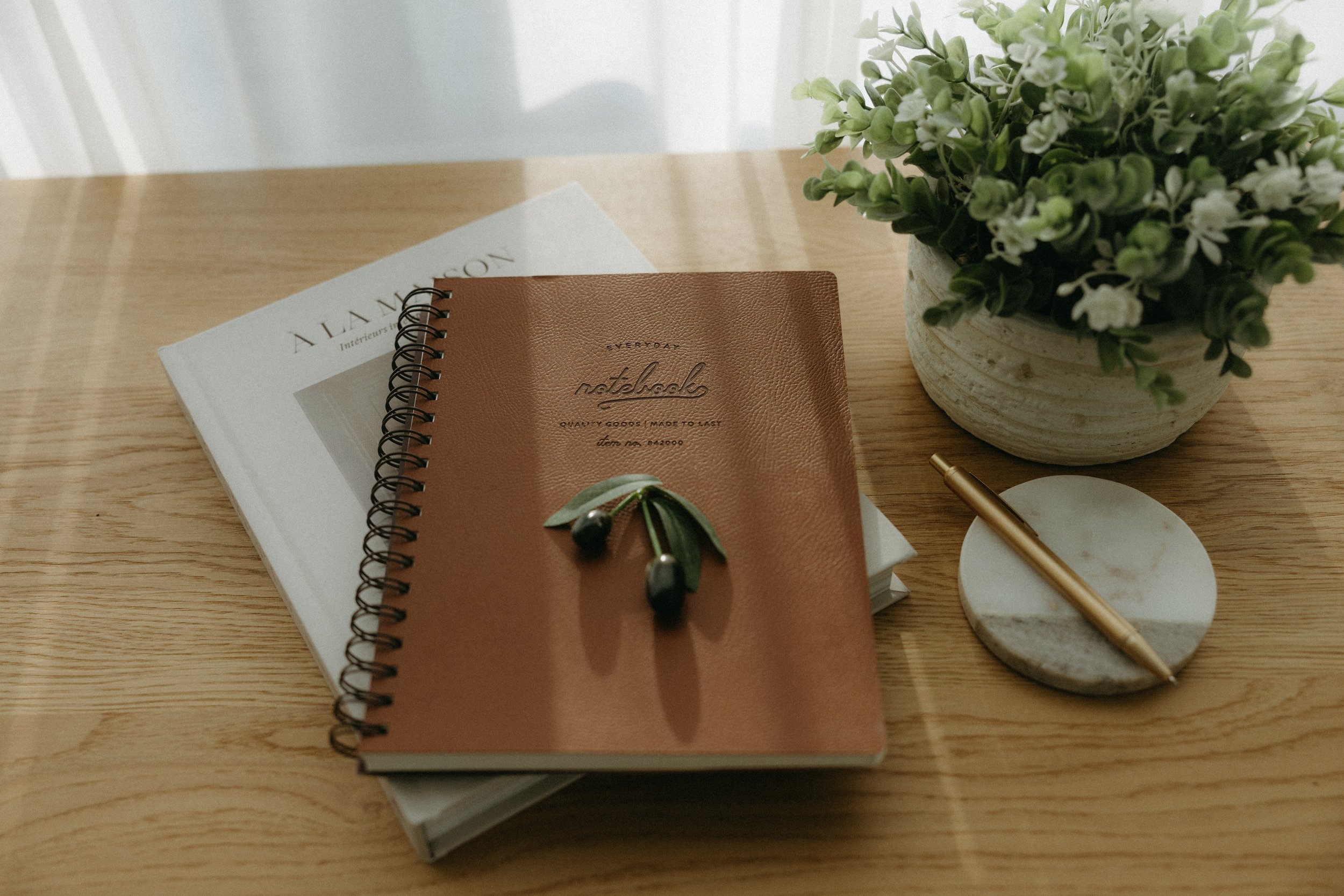 A wooden table with a white and beige notebook, a gold pen, and a potted green plant with small white flowers.