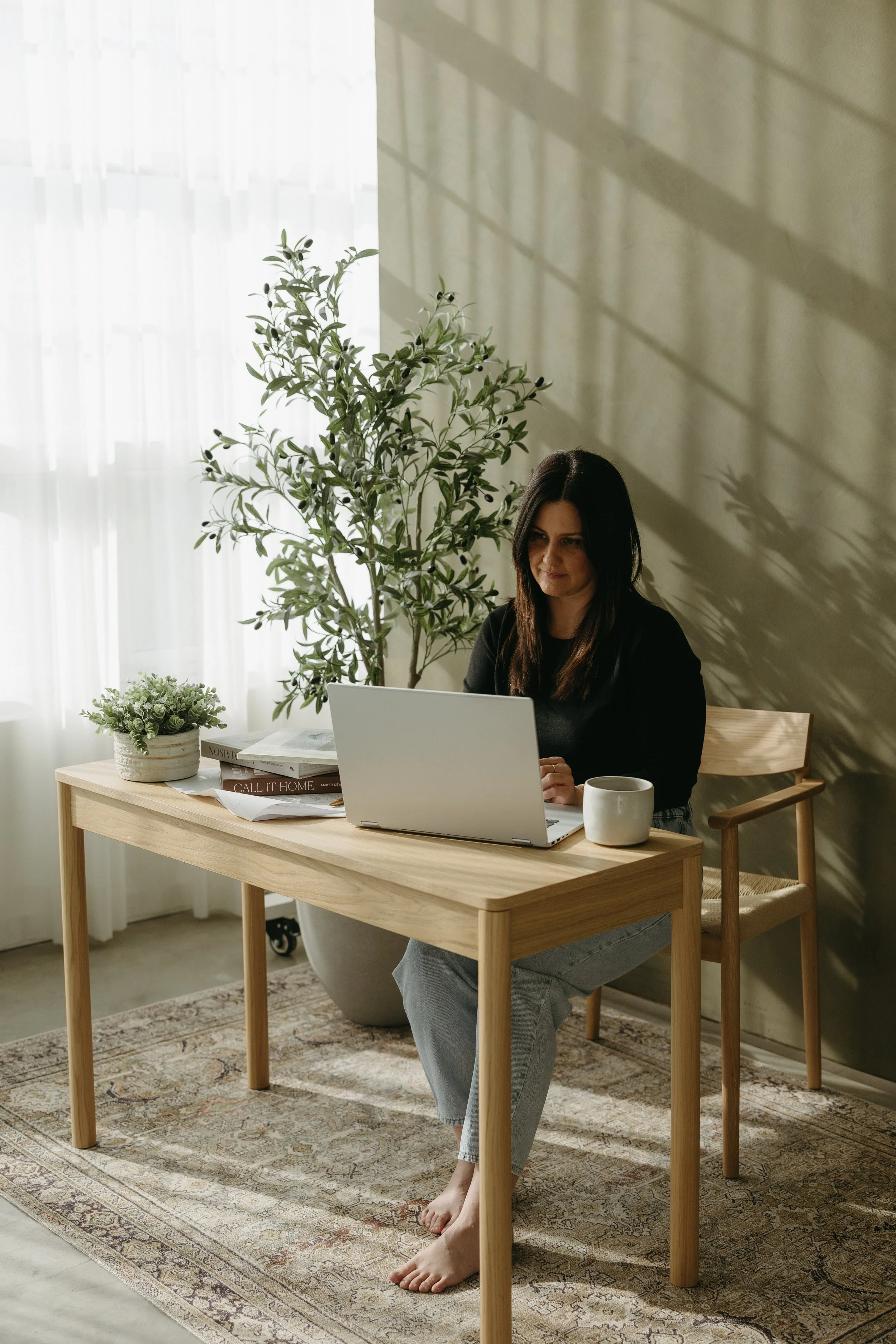 A woman with brown hair sitting at a wooden desk with a laptop, a coffee mug, and some books. She is in a room with light-colored walls, plants, and a large window with sheer curtains, casting shadows on the wall.
