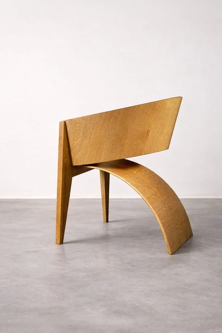 A wooden chair with a curved seat and slanted backrest, set on a plain light gray floor against a white wall.