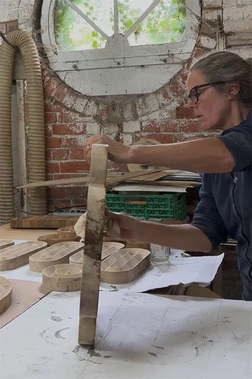 A woman with glasses and gray hair working with wood in a workshop. She is using a ruler or straight edge on a piece of wood, with several wooden molds or trays on the table. The workshop has a brick wall, a round window, and visible ventilation pipes.