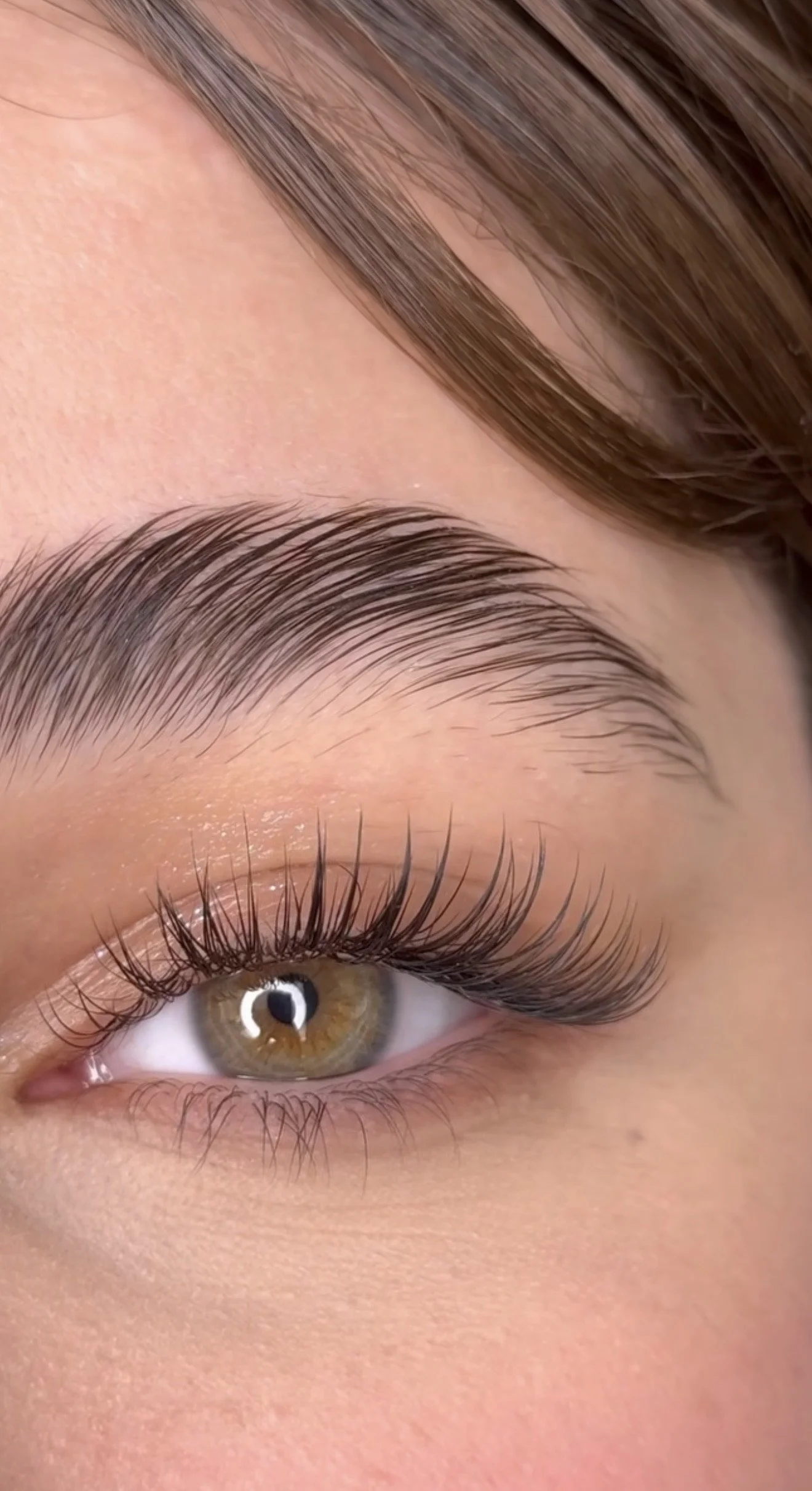 Close-up of a woman's eye with well-groomed eyebrows, long eyelashes, and light makeup, showing brown iris and detailed skin texture.