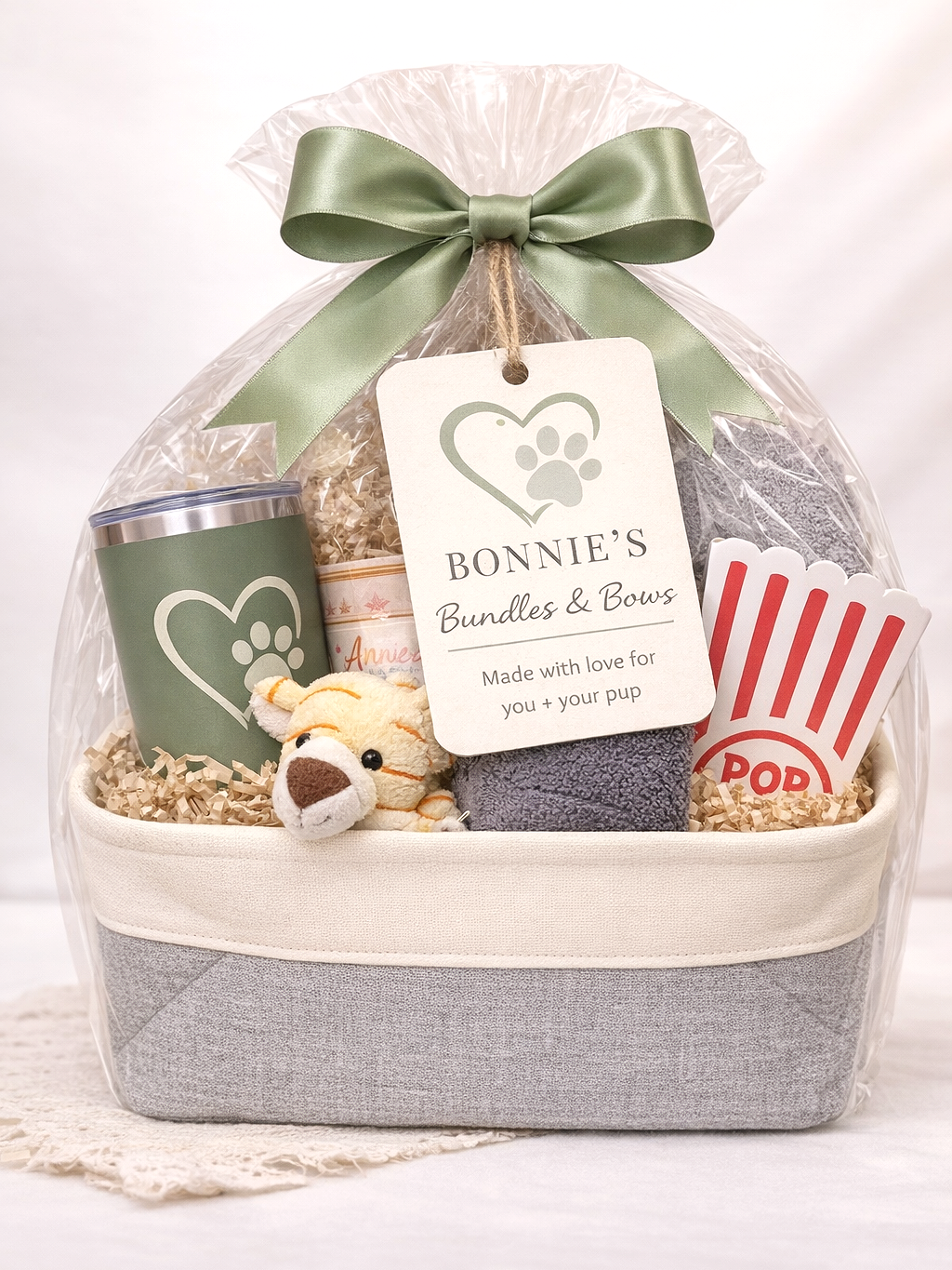 Gift basket wrapped in clear plastic with a green bow, containing a dog-themed tumbler, a small plush dog, a rolled towel, a canister, a popcorn box, and a towel, all with dog-themed packaging and label reading "Bonnie's Bundles & Bows".
