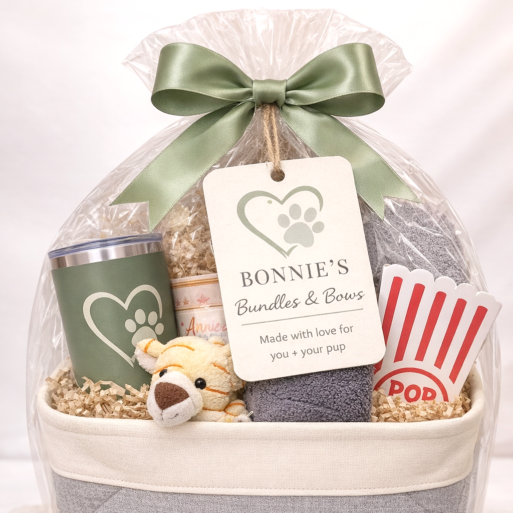 Gift basket wrapped in clear cellophane with a green ribbon bow, containing a tumbler with a paw print, a plush tiger toy, a container of dog treats, a small towel, and a popcorn container, with a tag reading 'Bonnie's Bundles & Bows, Made with love for you + your pup'.