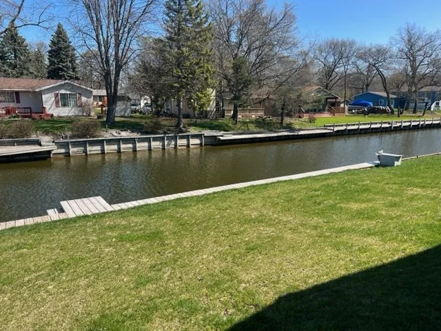 A backyard with a wooden dock extending over a canal leading to Lake Huron, green grass, trees, and a neighboring house.