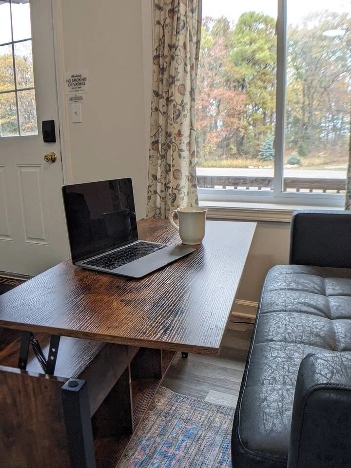 A wooden table with a laptop and a large mug on it, next to a leather couch, in front of a window at The Nook with floral curtains showing trees in Port Austin, MI.