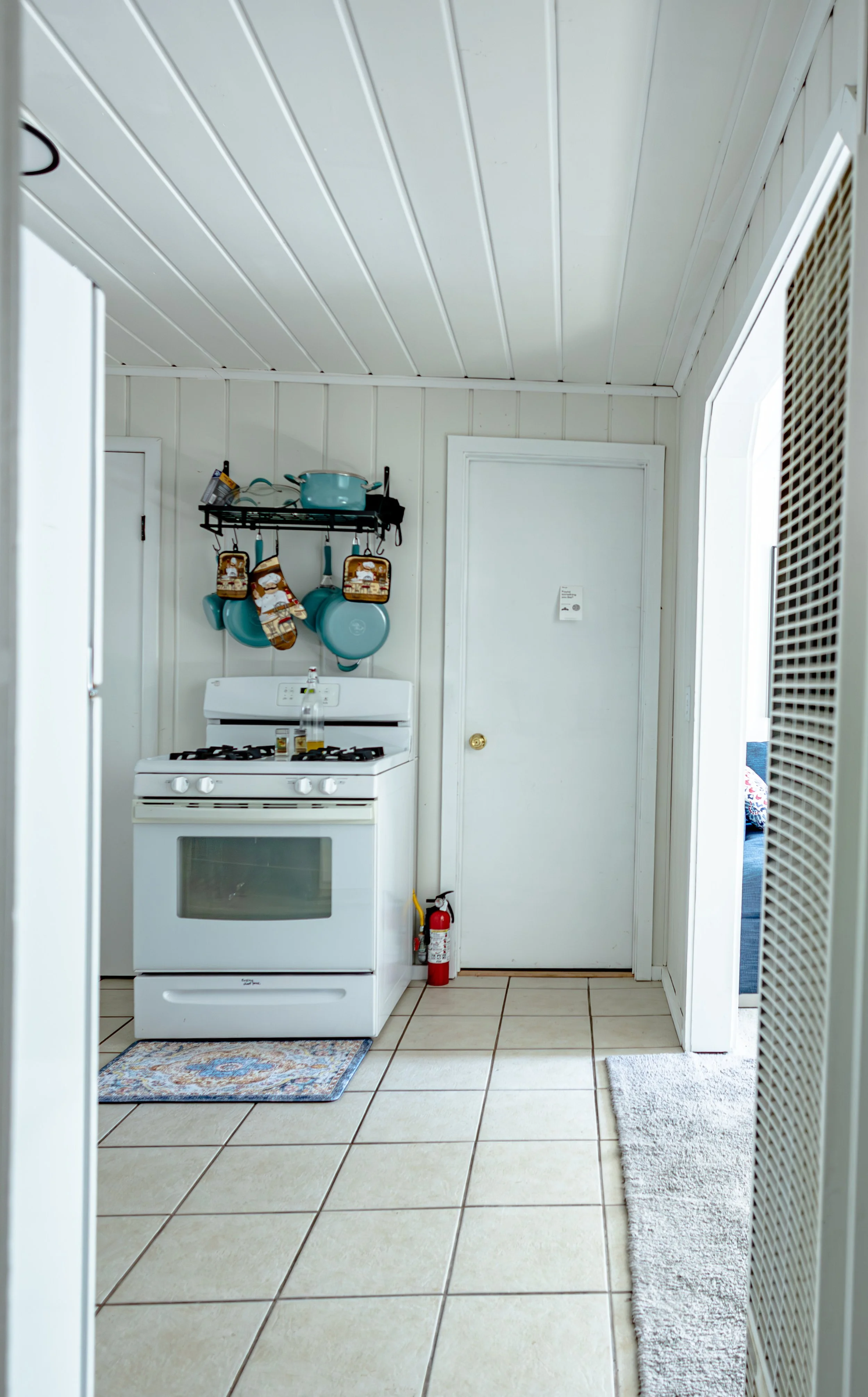 A small kitchen with a white stove, hanging blue pots and pans, a wall-mounted storage rack, tiled floor, and a door. A small fire extinguisher is on the floor near the door.