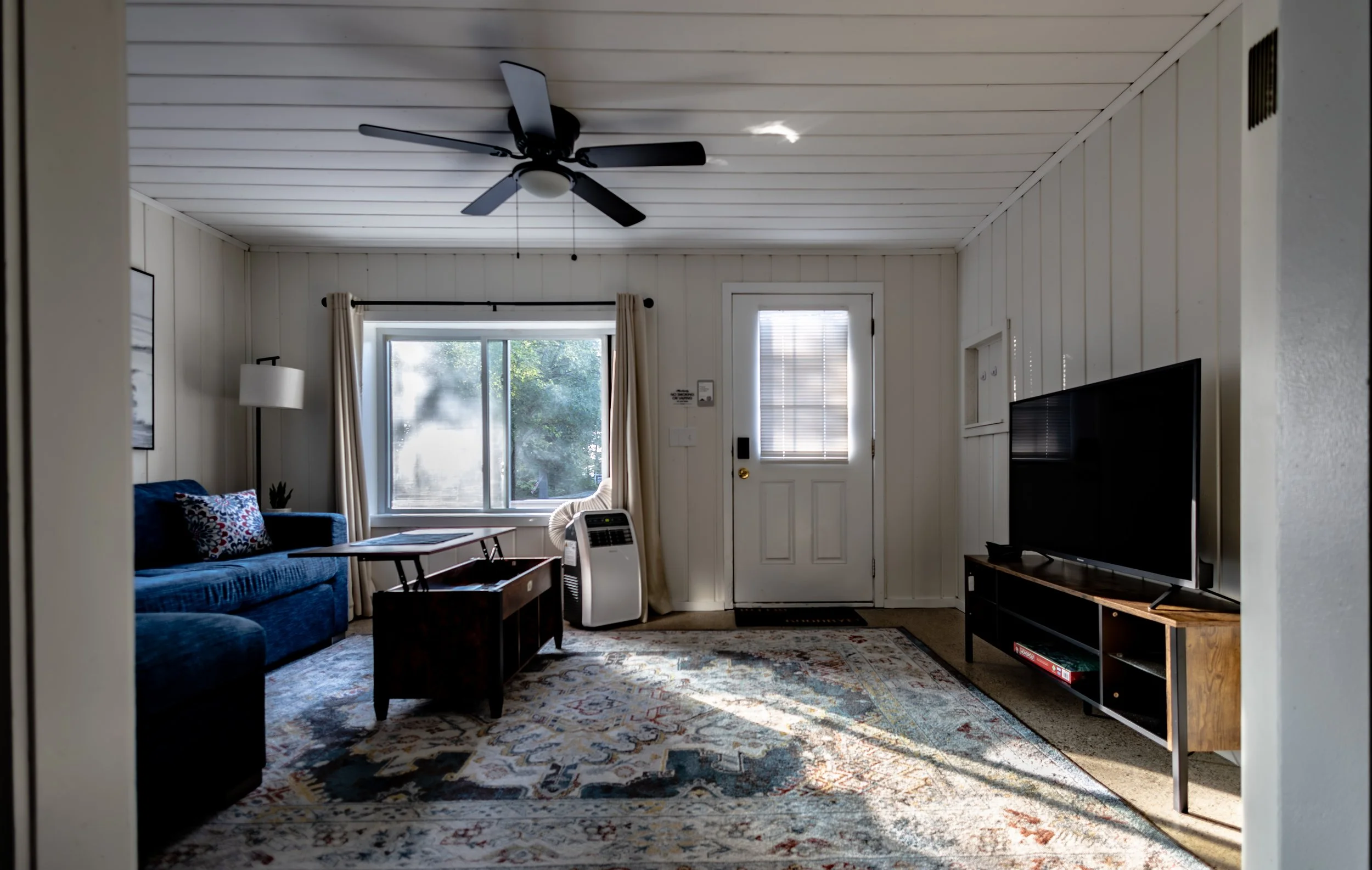 Living room with a blue sofa, a wooden coffee table, a large window with curtains, a TV on a wooden stand, a light-colored rug, a ceiling fan, and a door with a small window/blind.