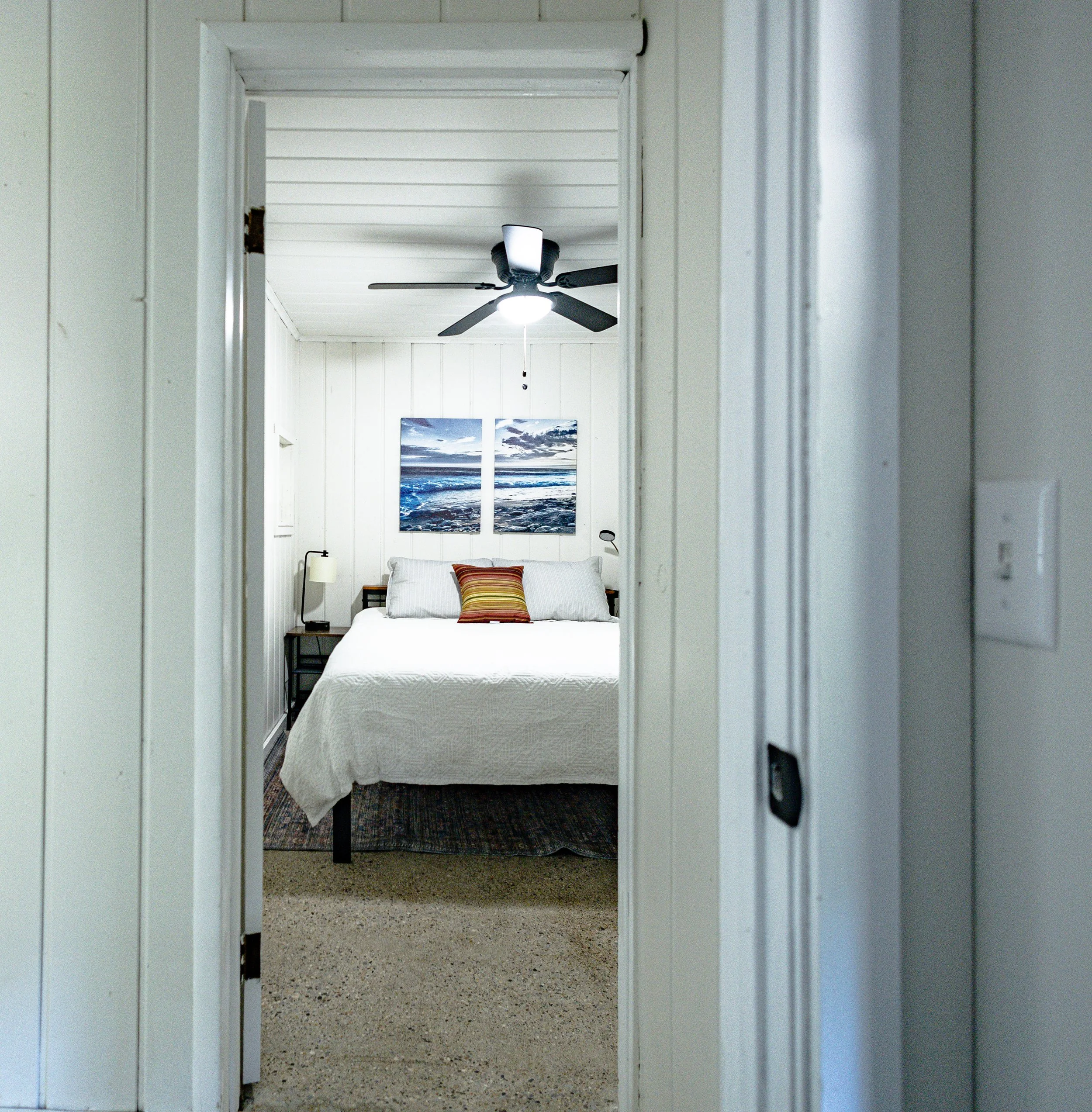 View through a door frame into a bedroom with white paneled walls, a bed with a white cover, a colorful striped pillow, two ocean art prints above the bed, a ceiling fan with a light, and bedside tables with lamps.