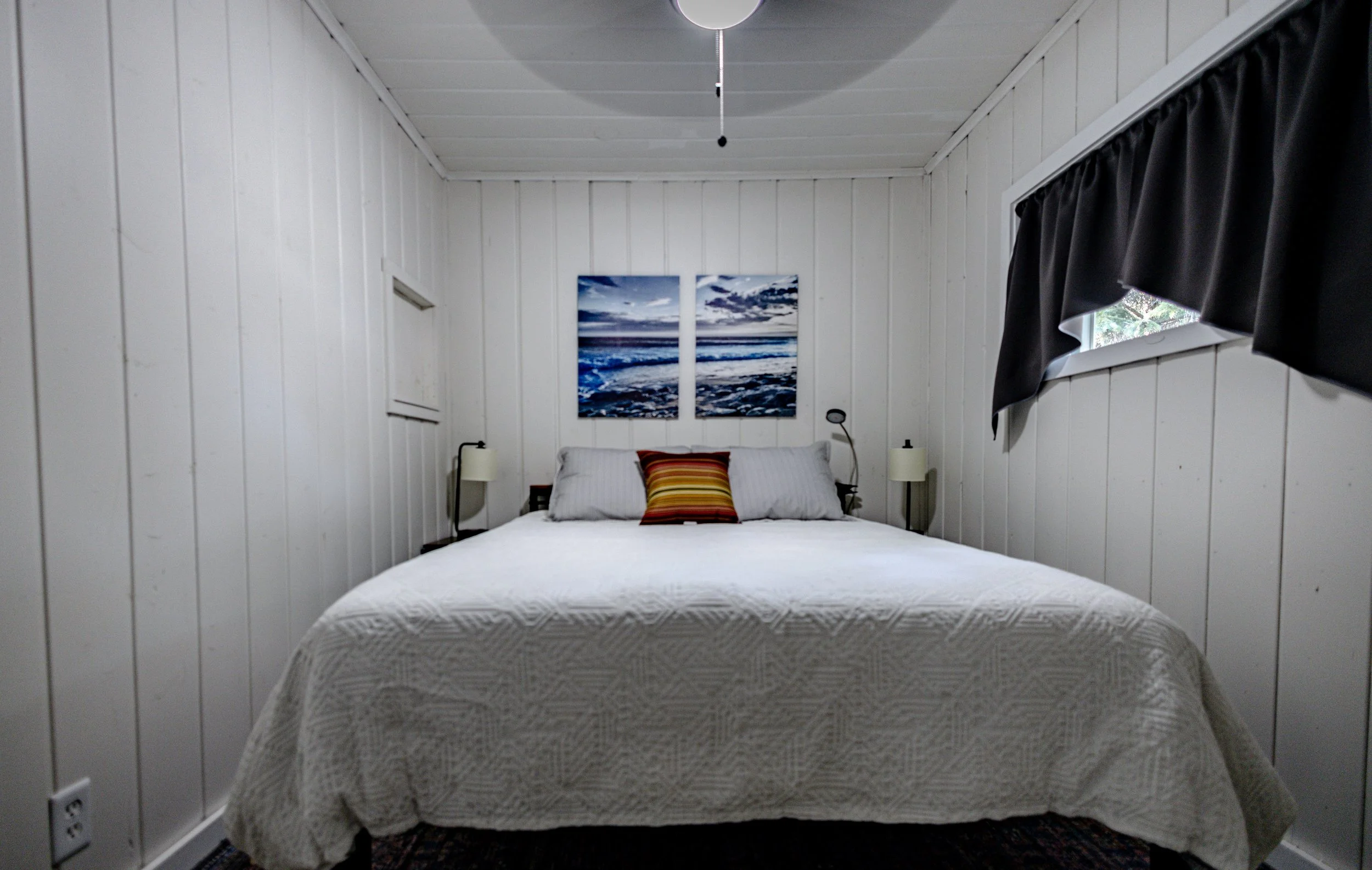 A cozy bedroom with white paneled walls, a bed with white bedding, two pillows, and a striped accent pillow. Above the bed are two seaside landscape paintings. Two black nightstands with lamps are on either side of the bed. There is a window with bla