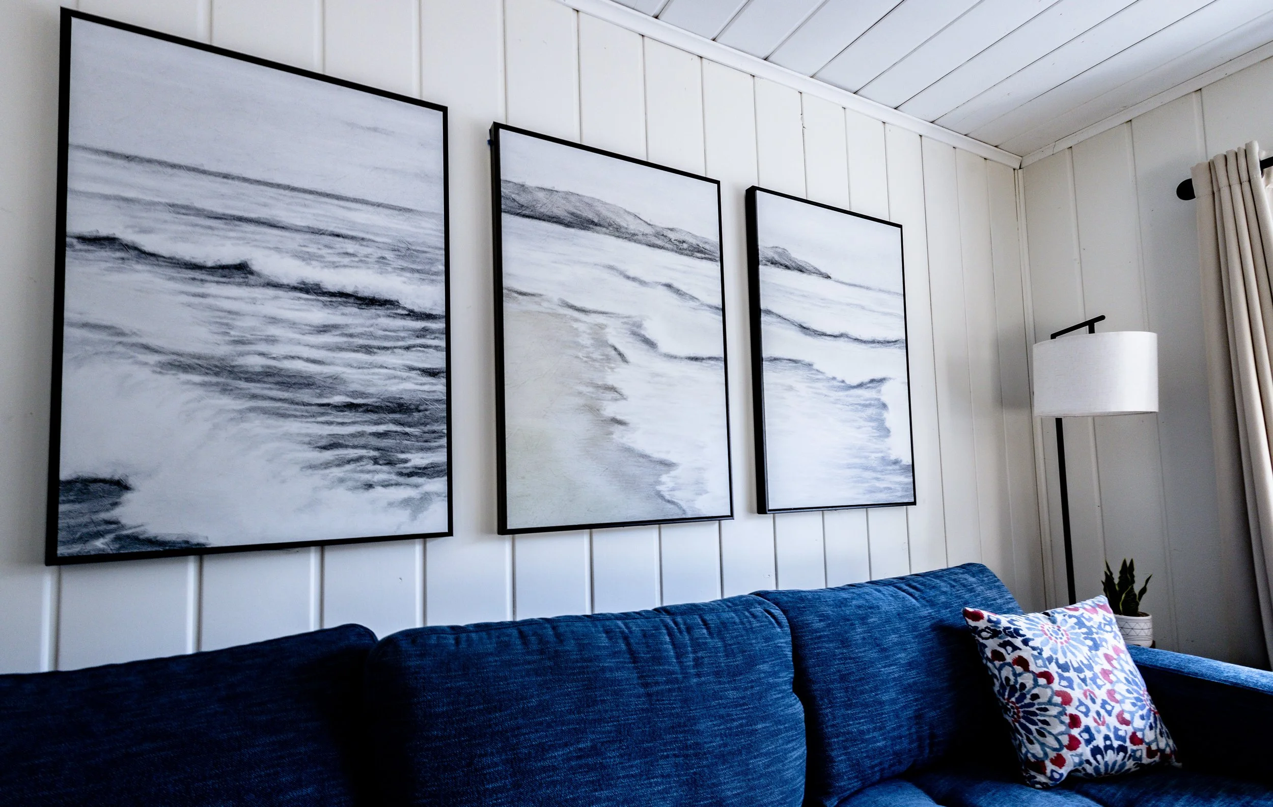 Living room with three black-framed abstract ocean-themed paintings on a white wall, a blue sofa with a patterned pillow, a white floor lamp, and a potted plant near a cream curtain.