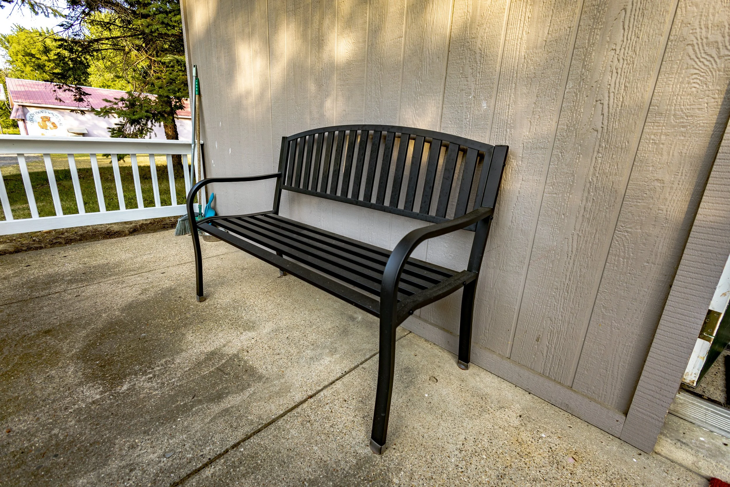 A black metal outdoor bench with a curved backrest and armrests. It is situated on a concrete porch next to a beige wooden wall, near a white railing, with trees and a building in the background.