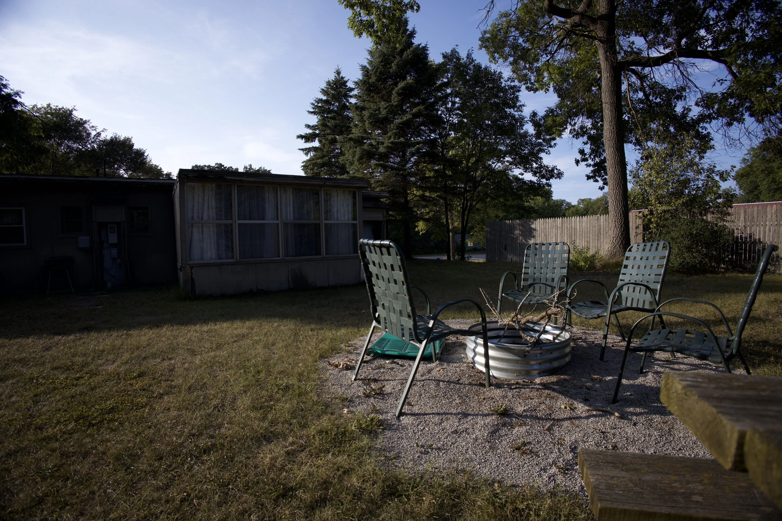Backyard with five green metal chairs arranged around a fire pit on gravel, surrounded by grass, trees, and a privacy fence, with a house in the background.