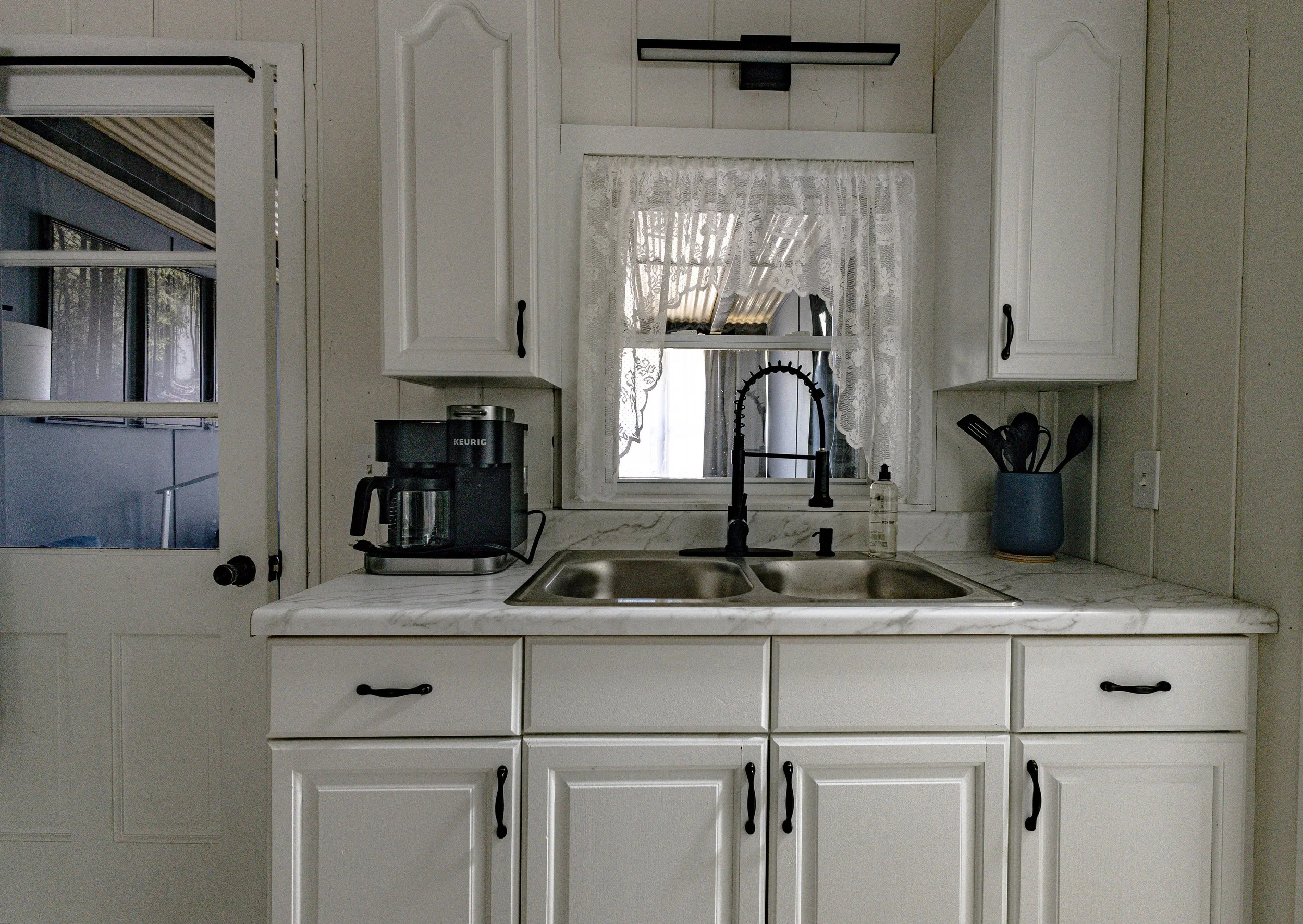 A kitchen with white cabinets and a marble countertop, featuring a black coffee Maker, a vintage style black faucet, a dish soap bottle, and a blue container holding utensils. There is a small window with lace curtains and a partially open door leadi
