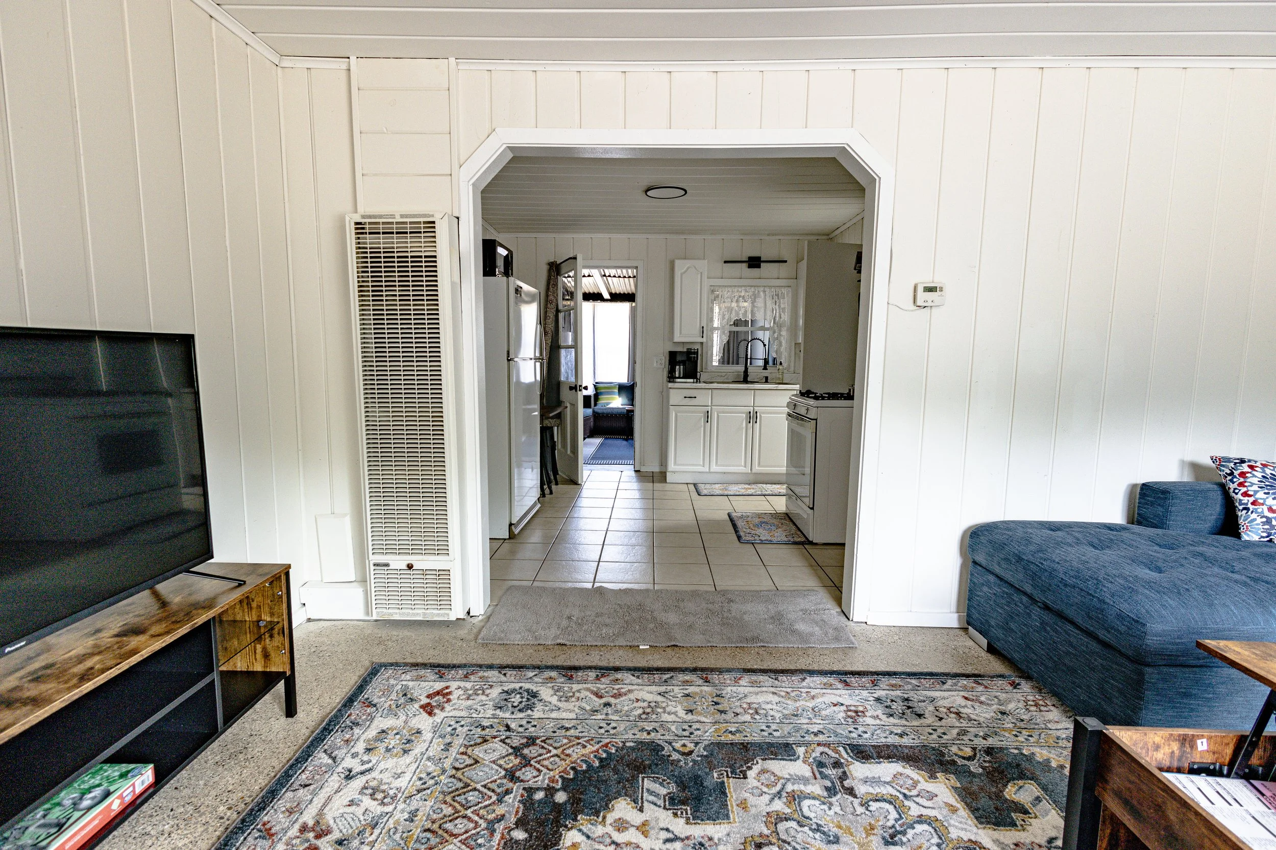 Living room view of a home interior with a television on a stand, a blue couch with a patterned pillow, a large rug, and a doorway leading to a kitchen with white cabinets, a refrigerator, and a stove.