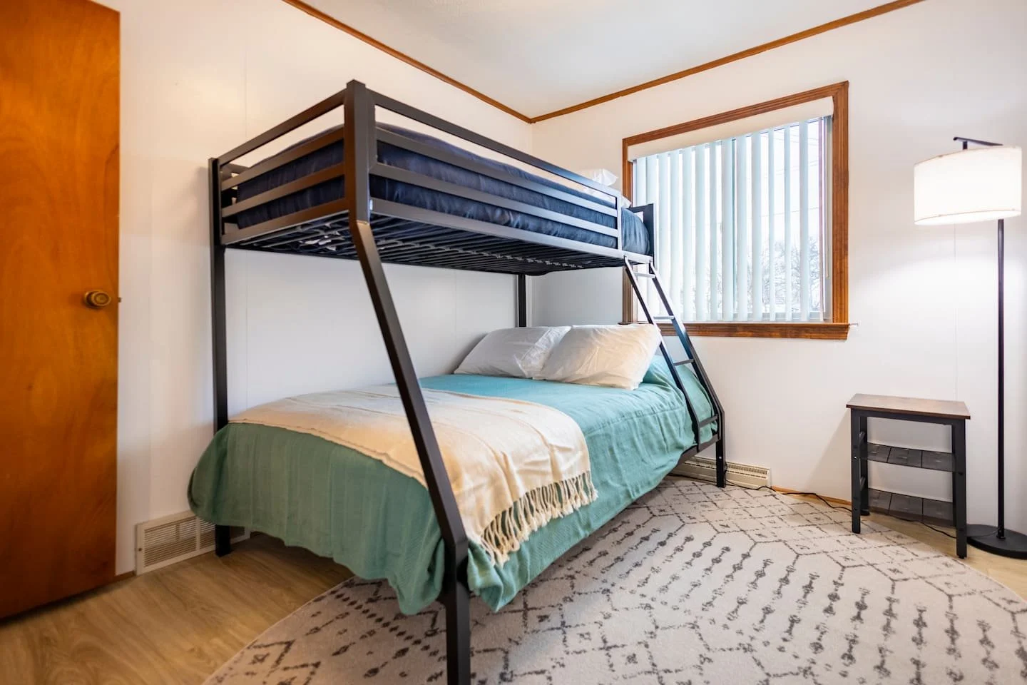 Bedroom with bunk beds over a twin bed, a window with vertical blinds, a wooden door, a rug, and a tall floor lamp.
