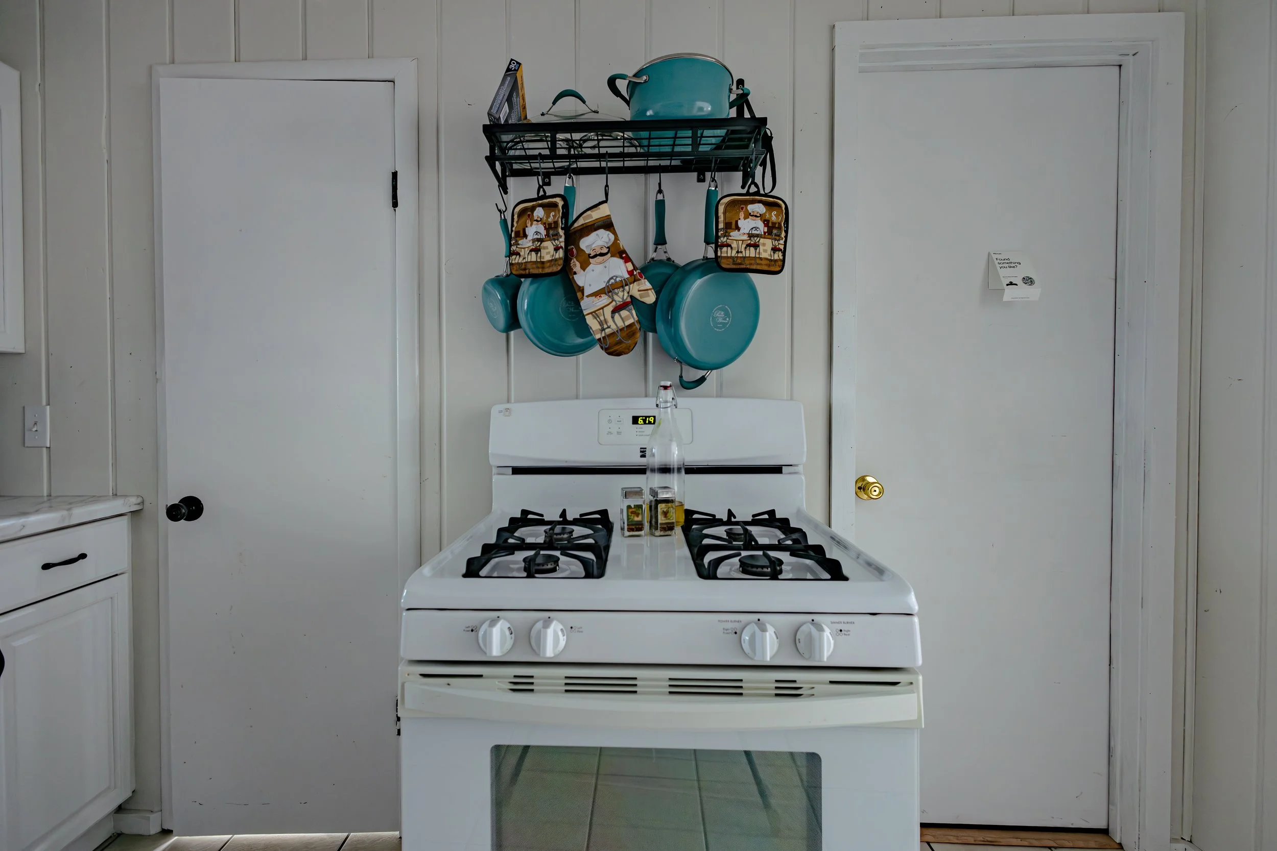 Kitchen with white stove, a rack above holding turquoise pots, pans, and kitchen accessories, with a bottle of water and condiments on the stove