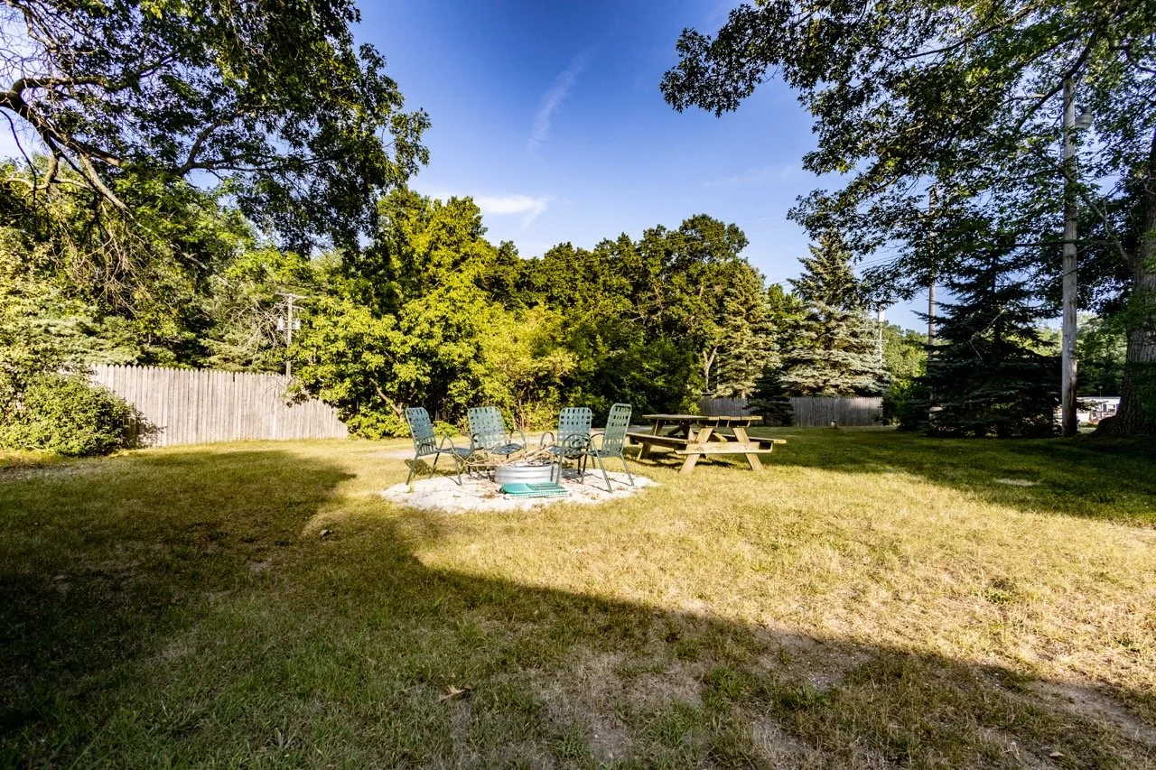Empty backyard with a fire pit surrounded by chairs, a picnic table, and trees underneath a clear blue sky.