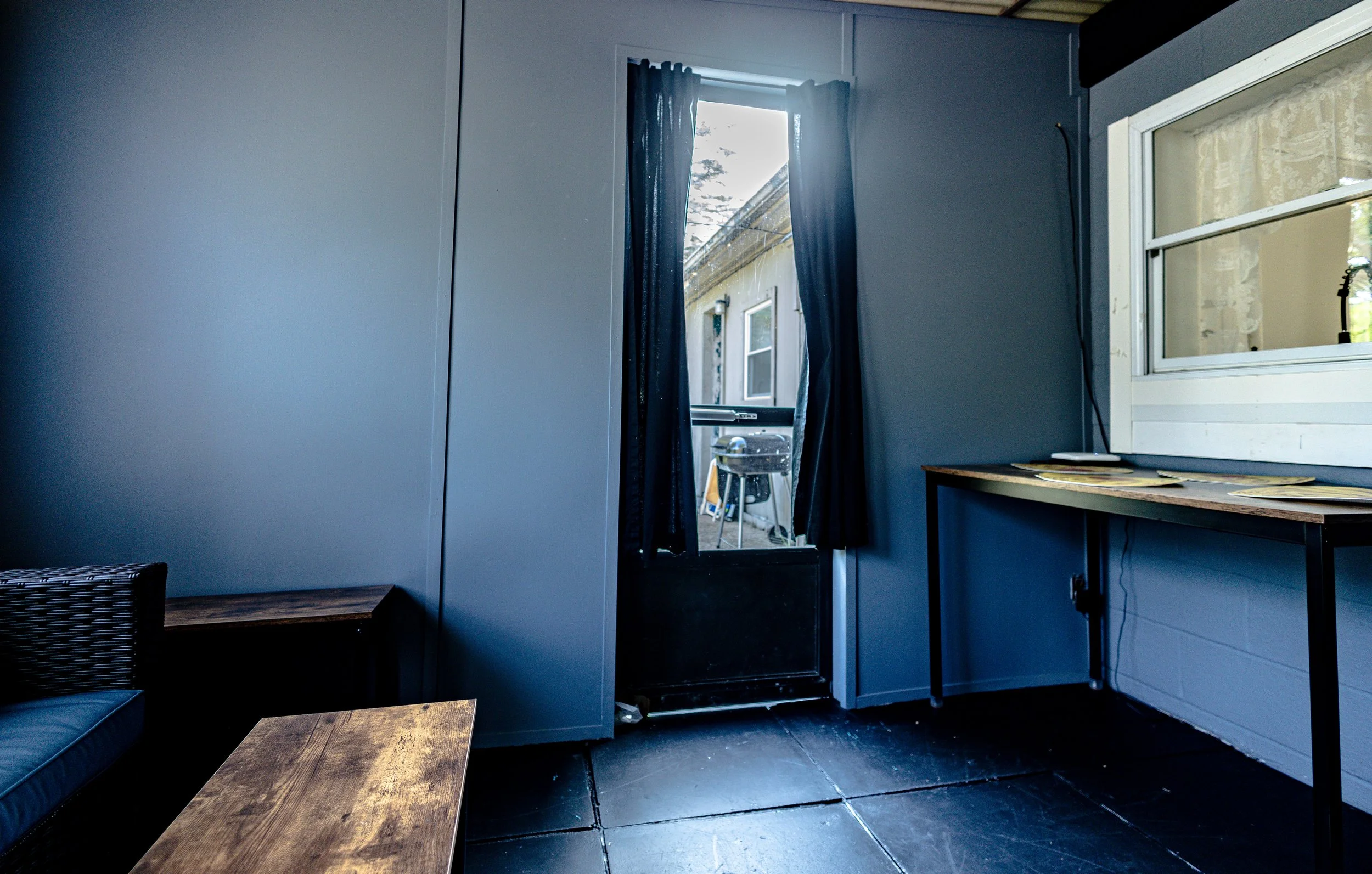 Interior of a porch or sunroom leading outside, with a door, a window, a table with papers, and a wicker chair.