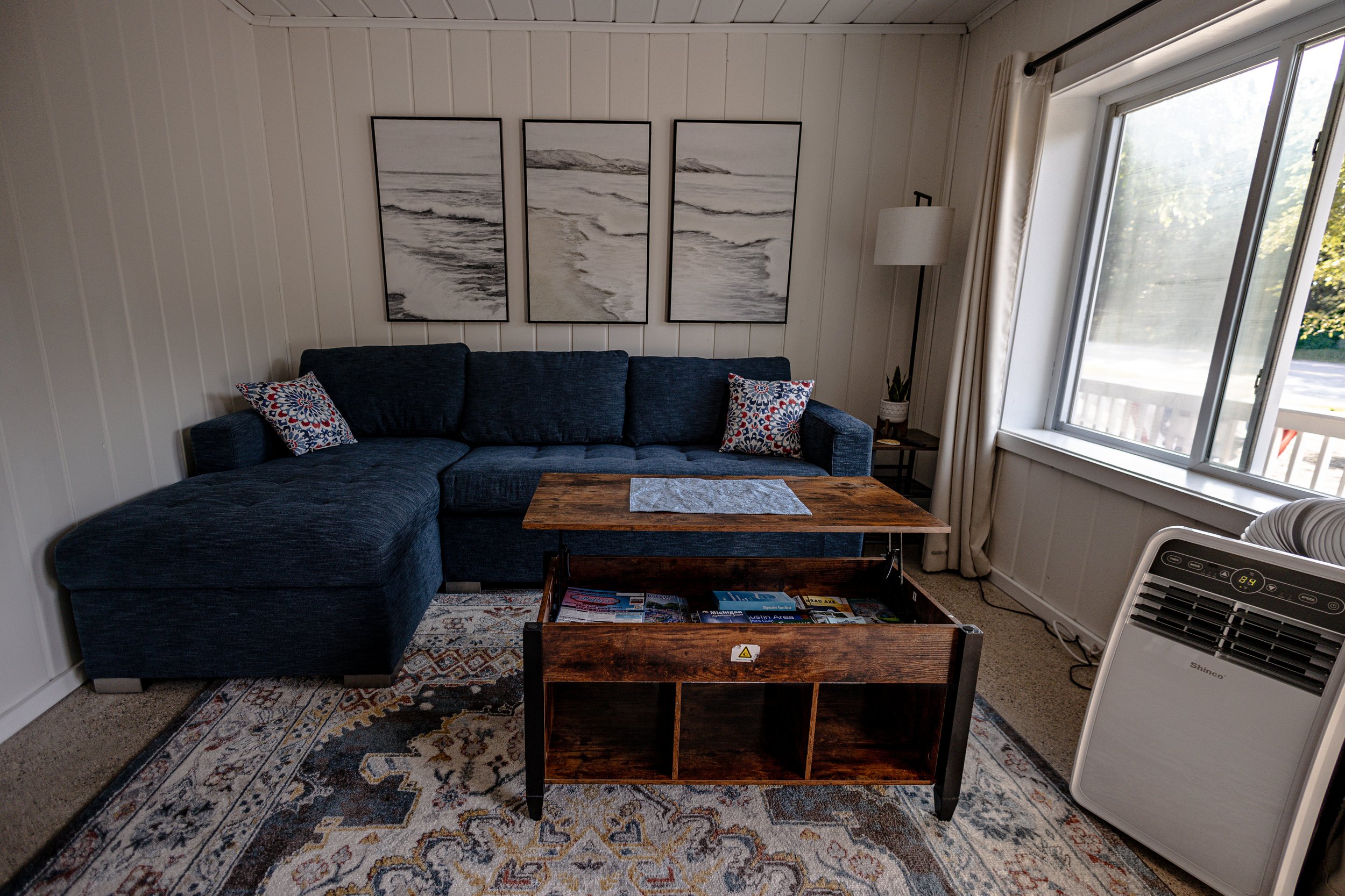 Living room with a dark blue sectional sofa, a wooden coffee table with shelves, a patterned area rug, a large window with curtains, a white portable air conditioner, and wall art depicting a beach scene.