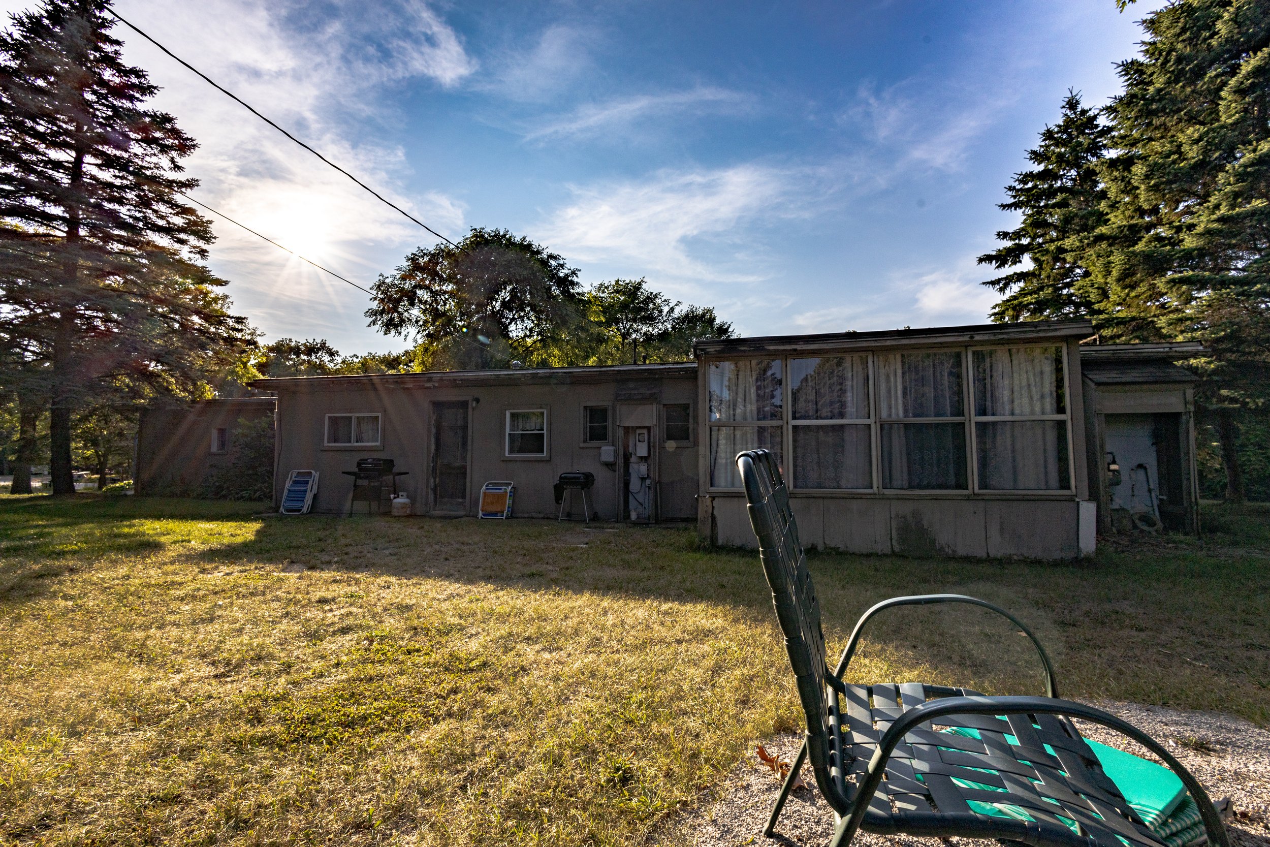 Backyard scene with a house, lawn chair, and outdoor items, with the sun shining through trees on a partly cloudy day.