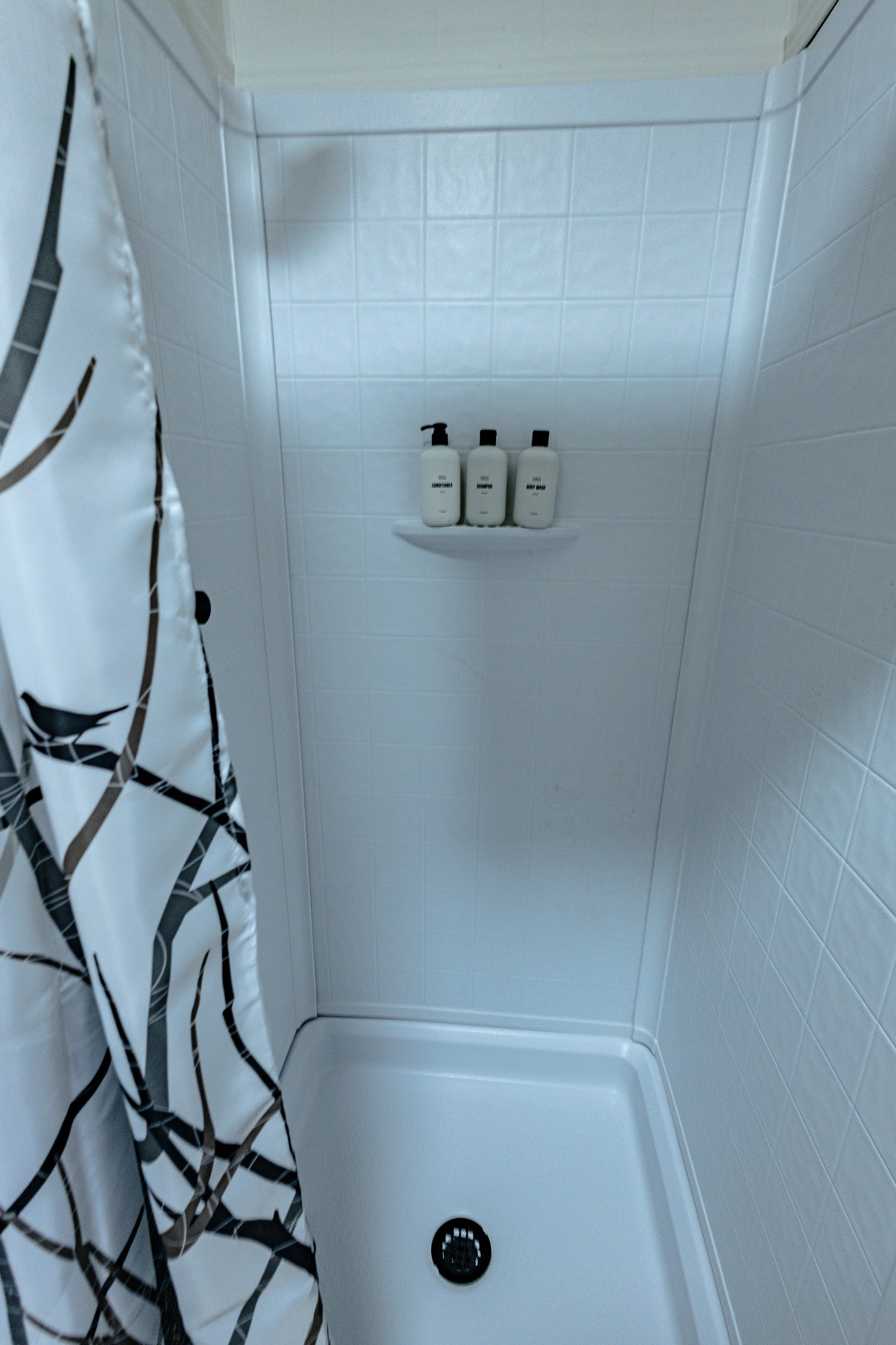 Empty shower stall with white tiled walls, a white shower base with a drain, and three bottles of shampoo and body wash on a small shelf, partially covered by a black and white patterned shower curtain