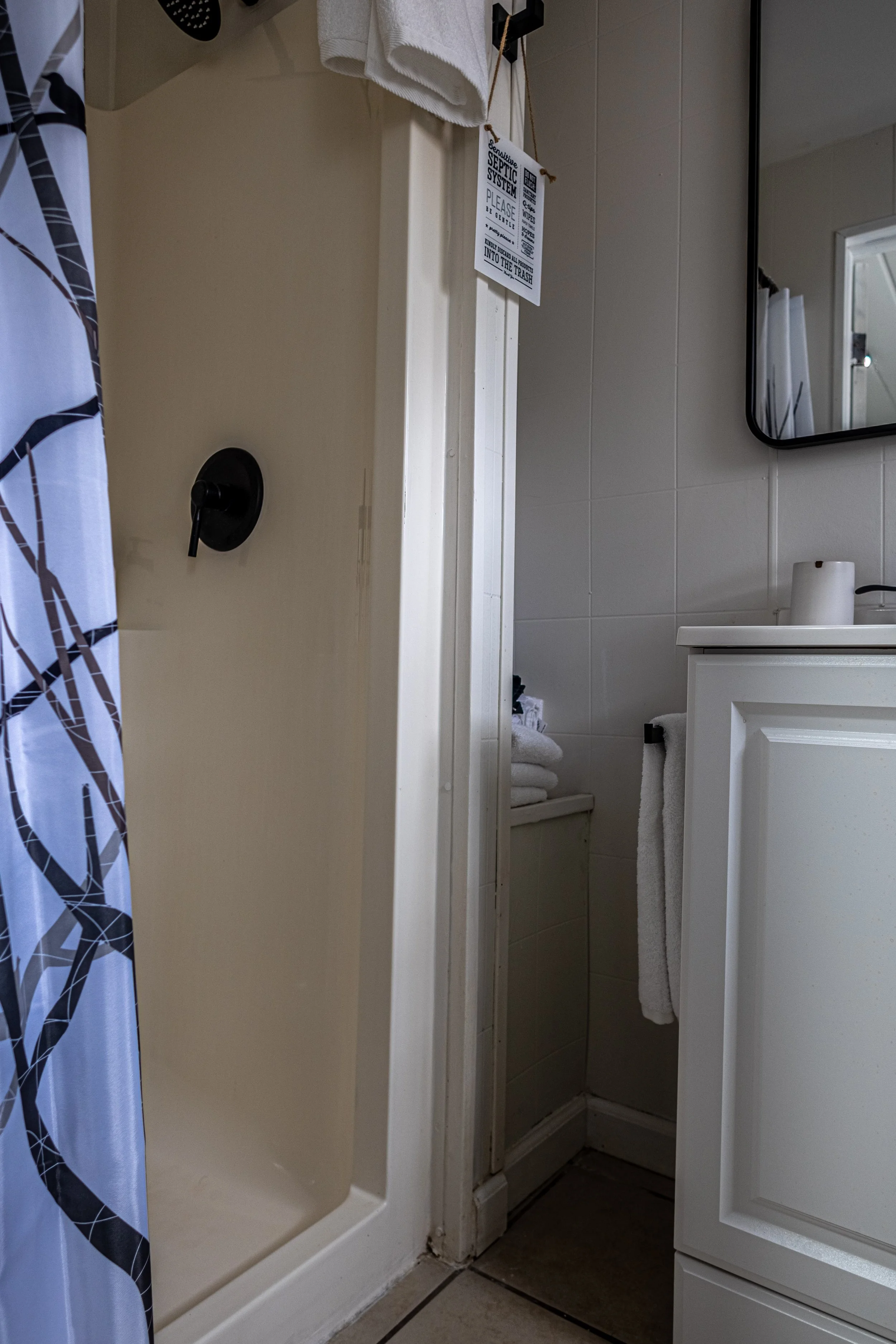 A small bathroom with a shower stall, a white cabinet with a towel hanging, a mirror, and a soap dispenser. Towel and toiletries are on the cabinet, and there is a tag hanging from a hook.