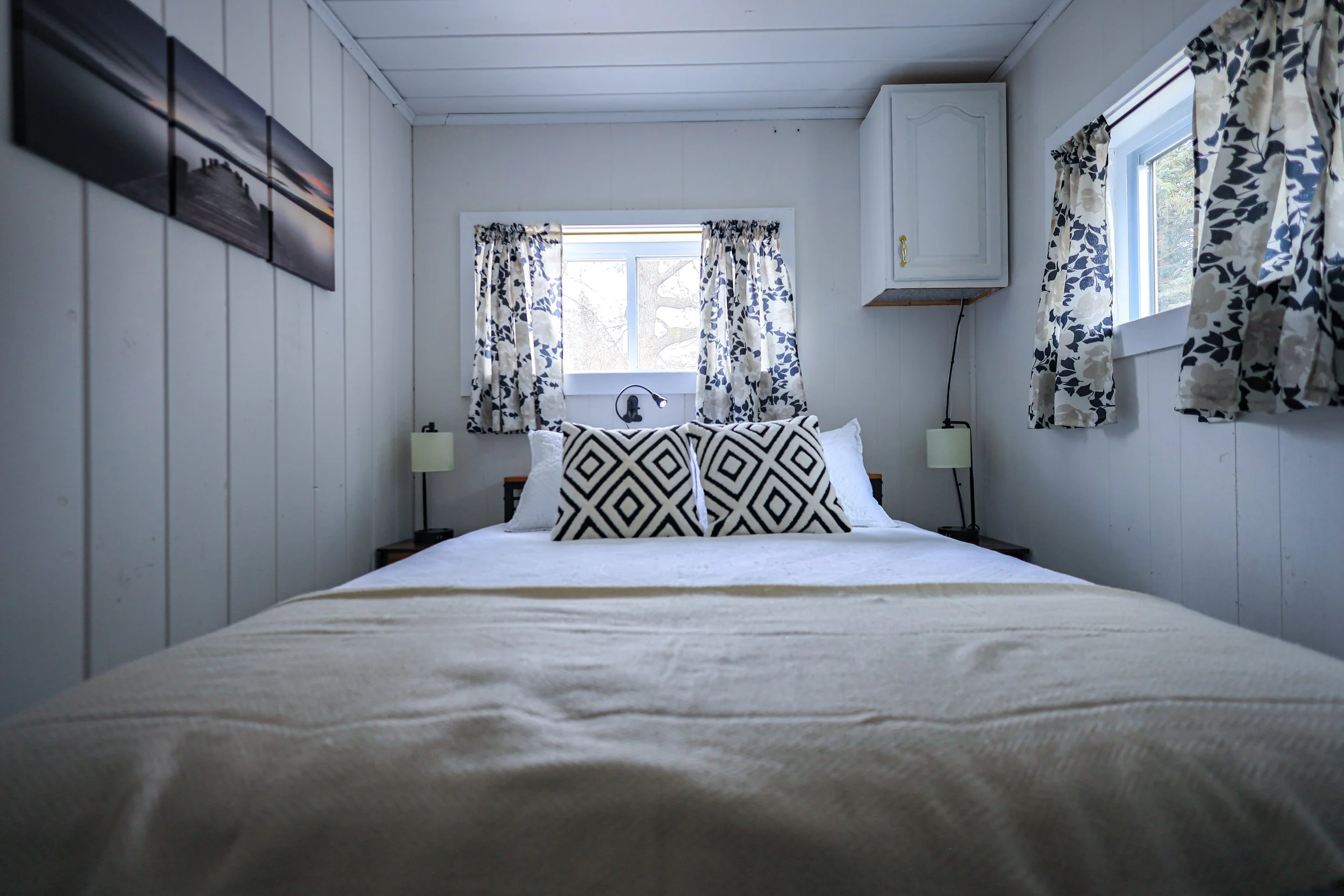 A cozy bedroom with white panel walls, a large bed with white bedding and black-and-white geometric pillows, two nightstands with lamps, and three windows with patterned curtains letting in natural light.