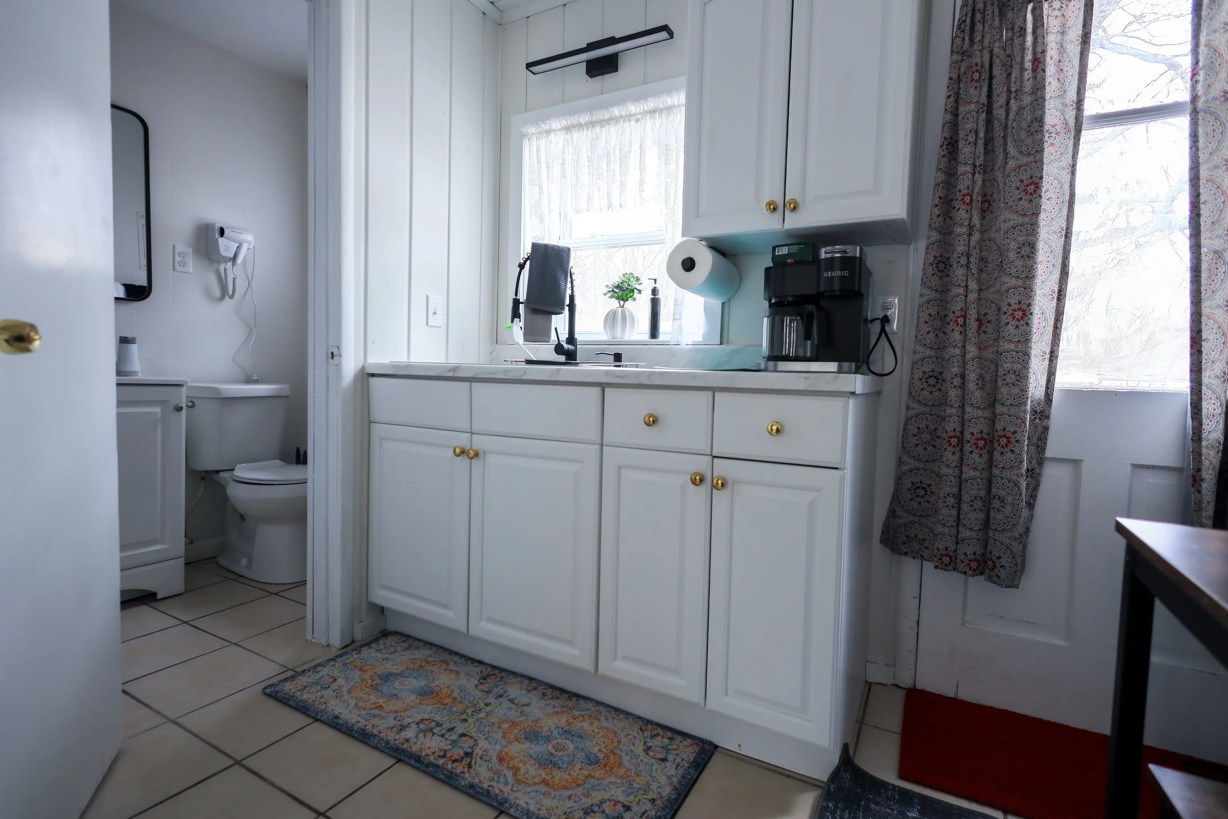White kitchen with a window, cabinets with gold knobs, countertop with a coffee maker, paper towel, and a small plant; adjacent bathroom with toilet and wall-mounted hairdryer; patterned curtains and rugs.