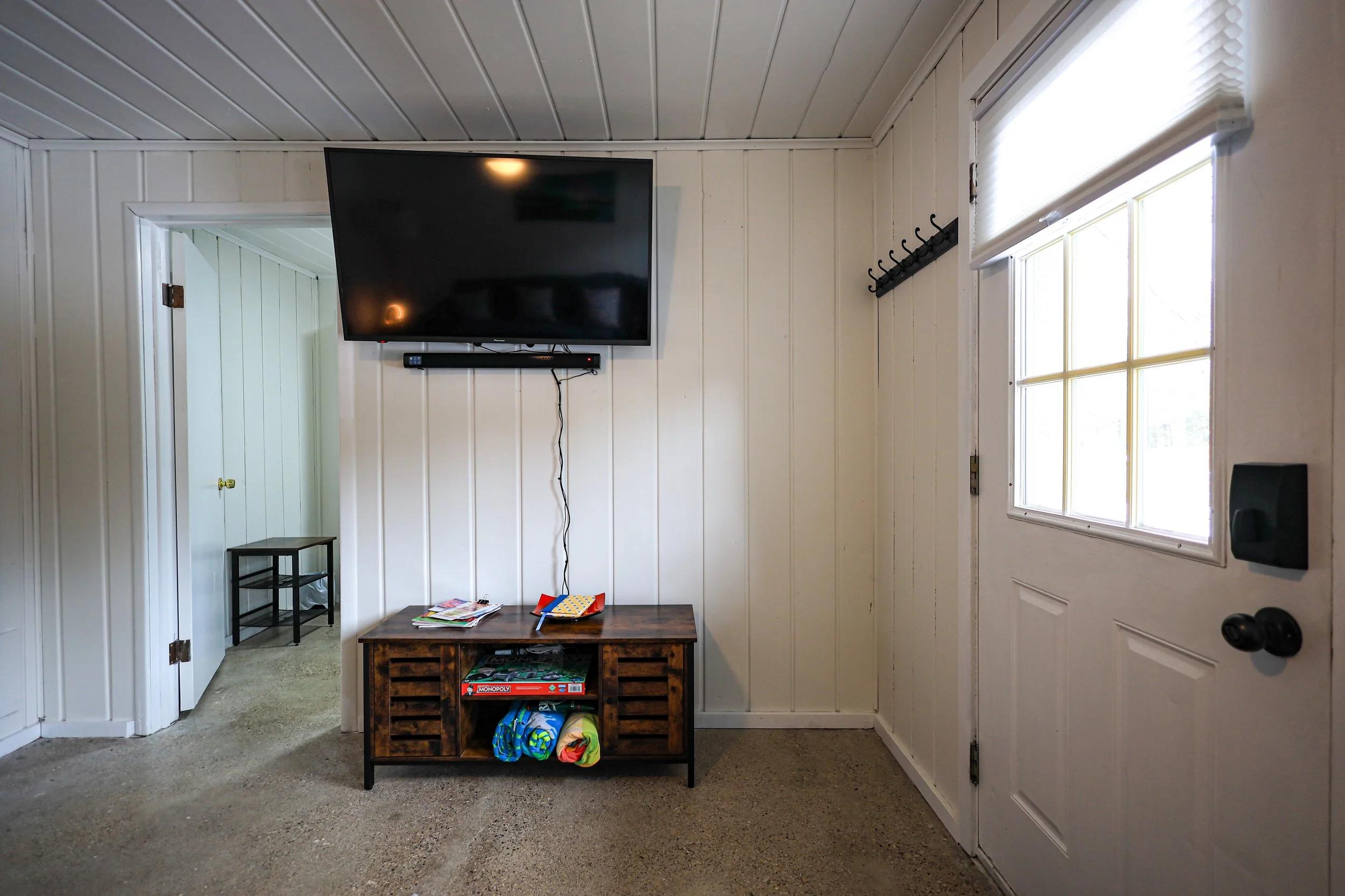 An interior living space with white paneled walls and a sloped ceiling. A large flat-screen TV is mounted on the wall above a wooden media console holding magazines and colorful rolled-up blankets. There is a doorway to a small room with a black tabl