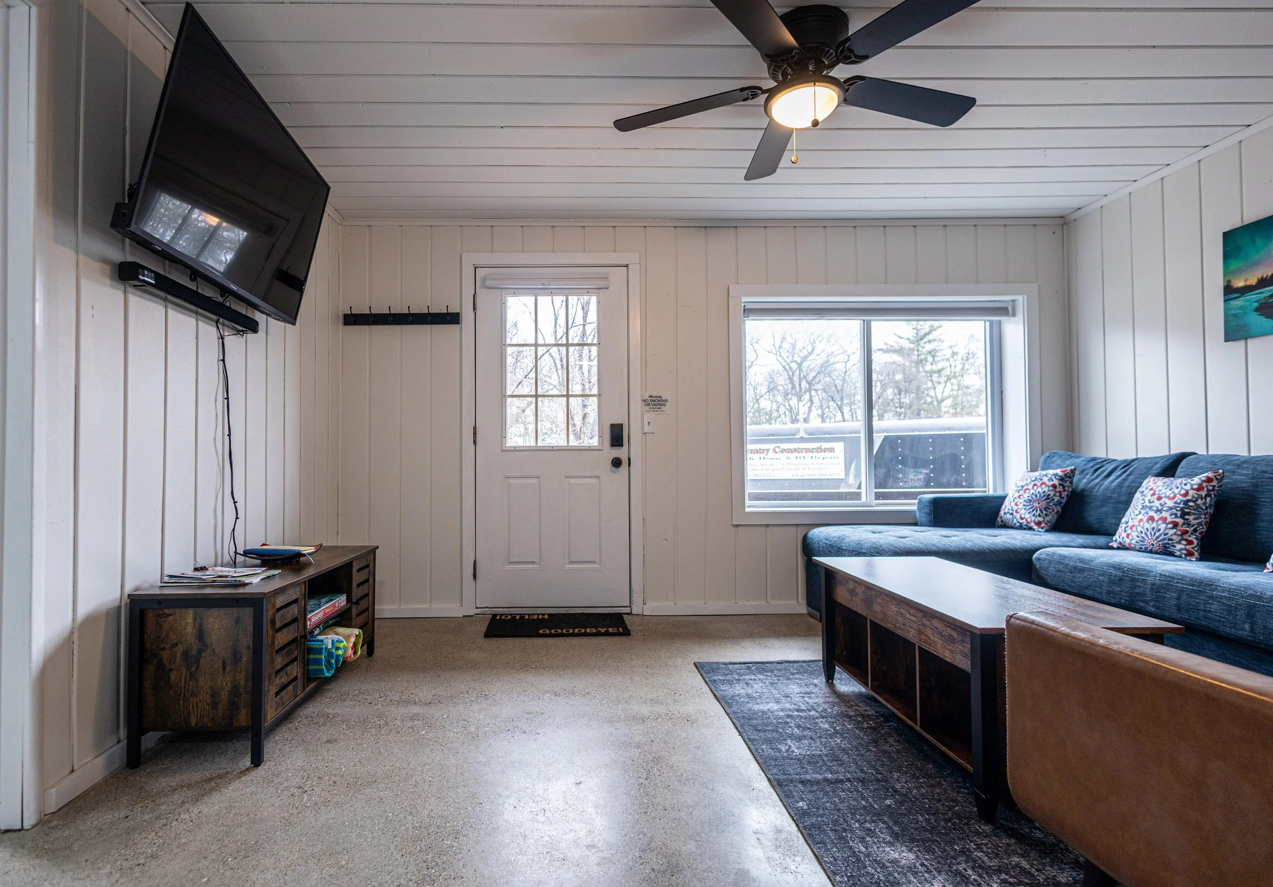 Living room with white paneled walls, a ceiling fan, a wall-mounted TV, a window, a blue sectional sofa with patterned pillows, a wooden coffee table, a brown armchair, and a door with a window.