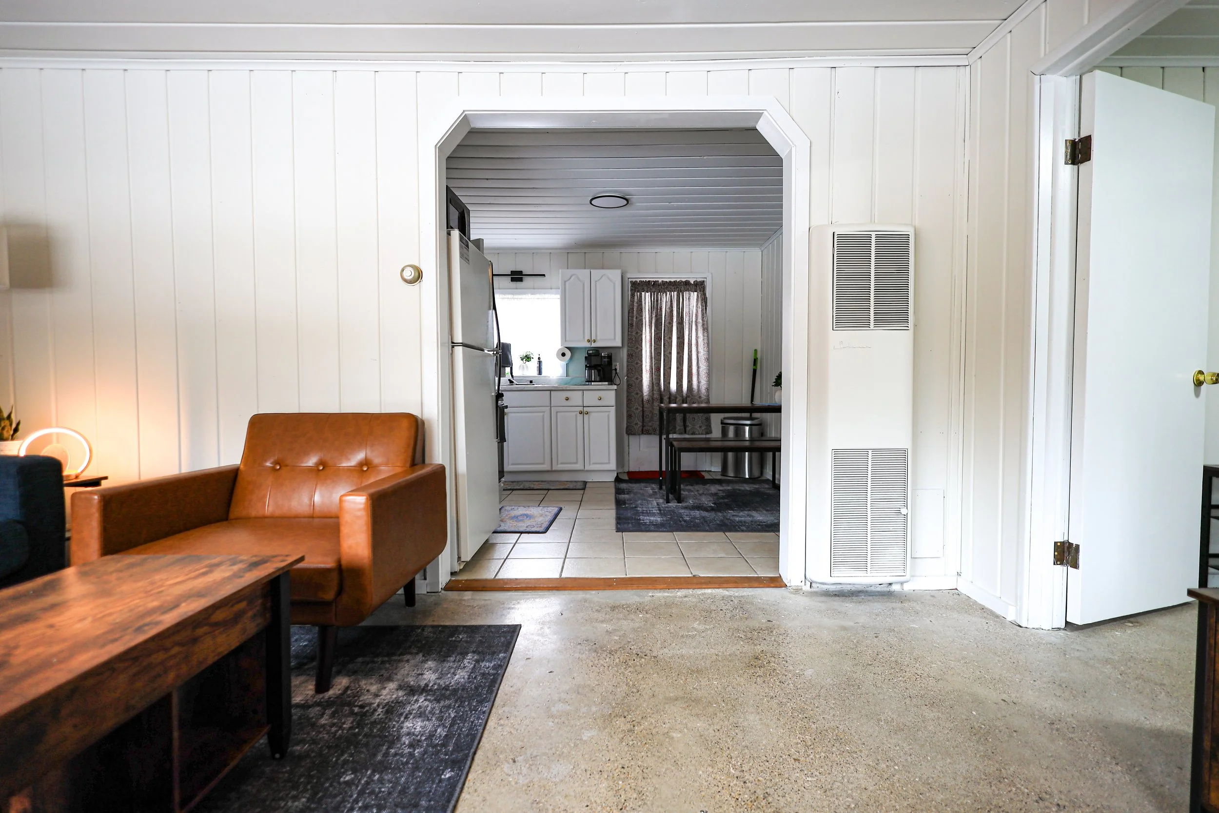 View of a living room with front entry to a kitchen, white paneled walls, brown leather armchair, side table, black rug, tiled kitchen floor, white cabinetry, window with curtains, small dining area with table and chair, and an open white door to the