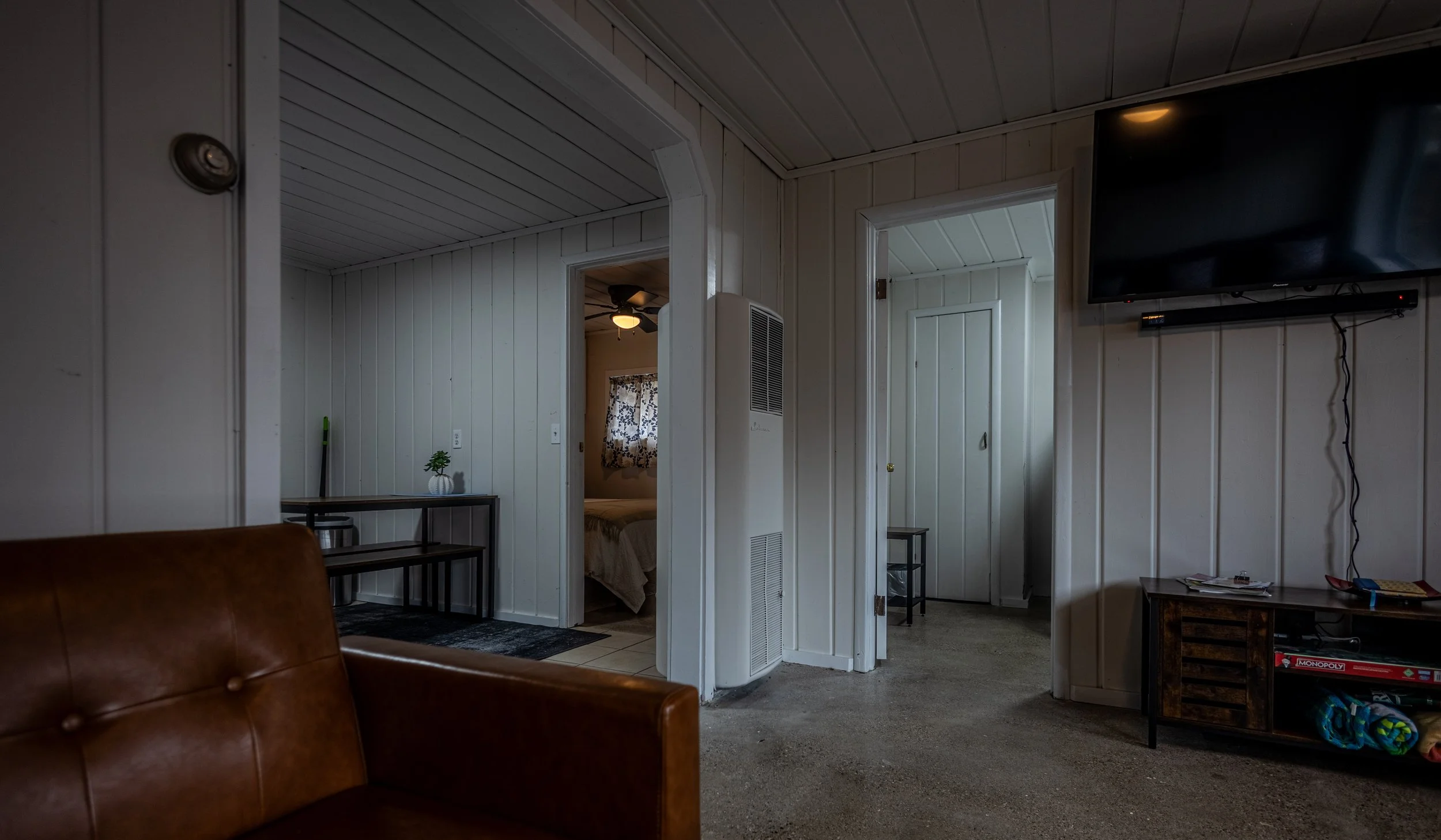 Living room with white paneled walls, a wall-mounted flat-screen TV, and an opening leading to a bedroom with a bed and window with patterned curtains.